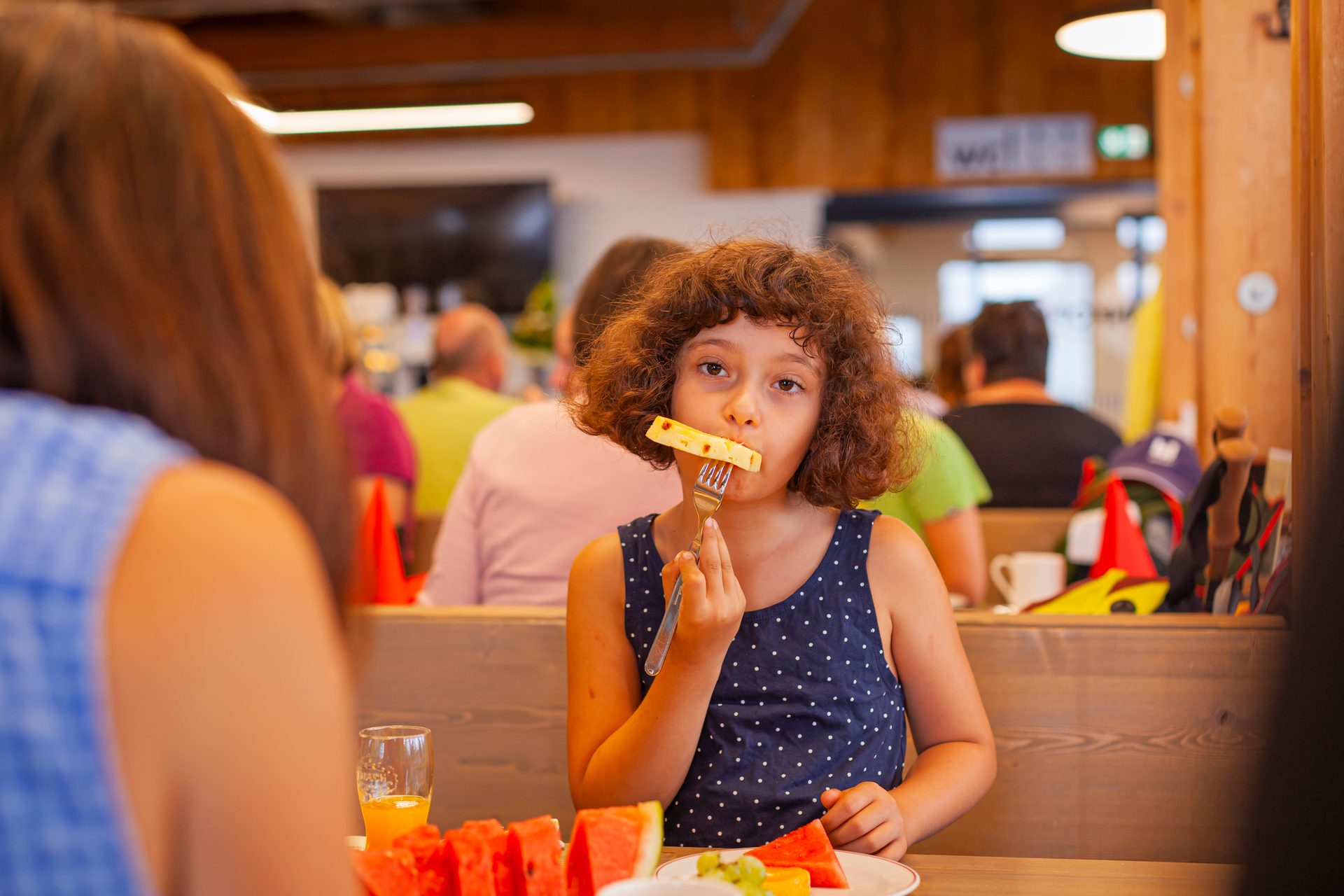 A young girl with curly hair eats fruit from a fork at a table with watermelon and orange juice.