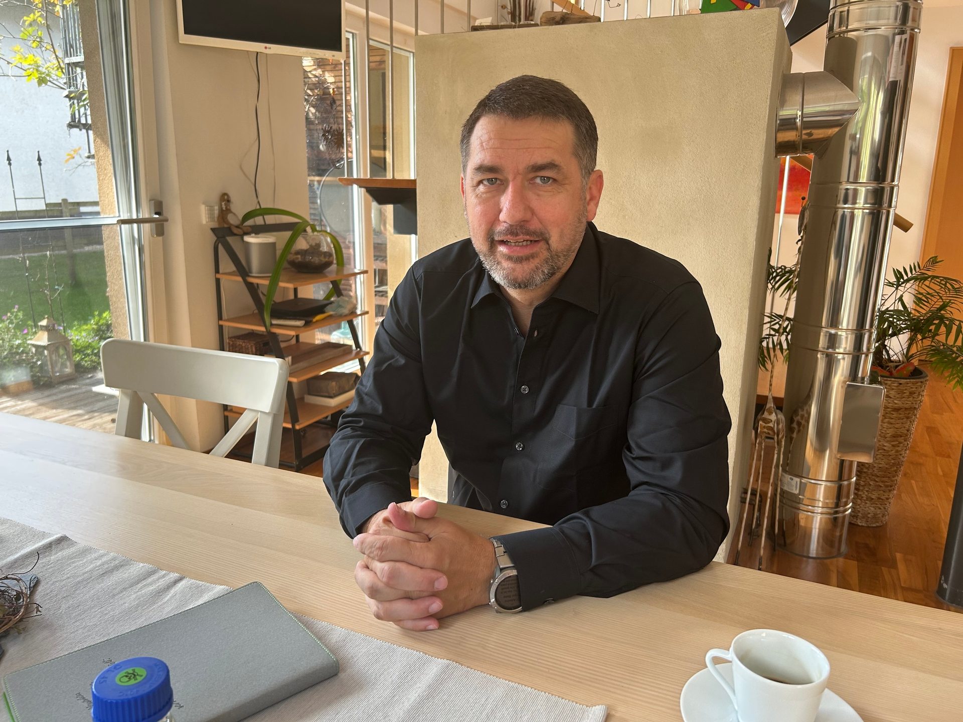 A man in a black shirt sits at a table, looking at the camera.