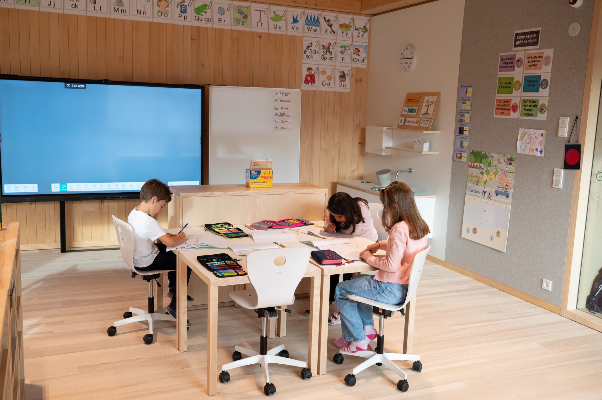 Three children studying in a bright, modern classroom with a digital whiteboard.