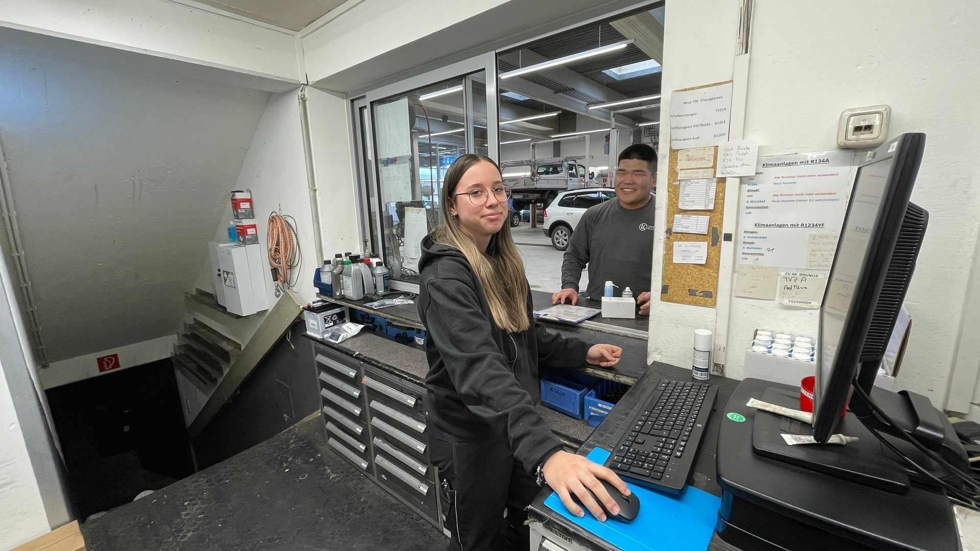 Two smiling auto service staff, a woman and a man, are seen at the counter of a garage.