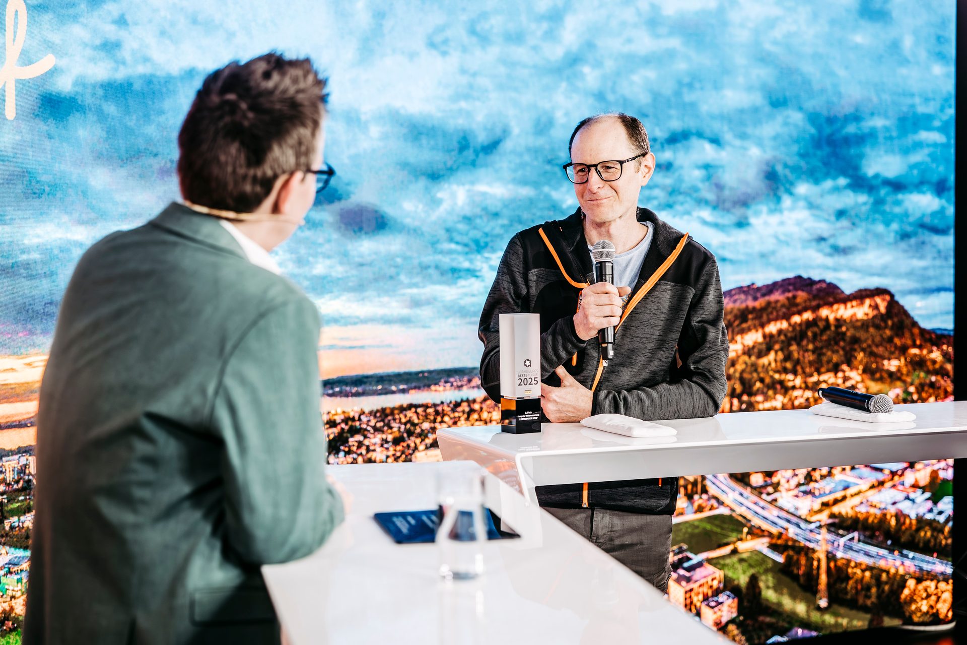A man speaks into a microphone, another person listens, with an award on a table.