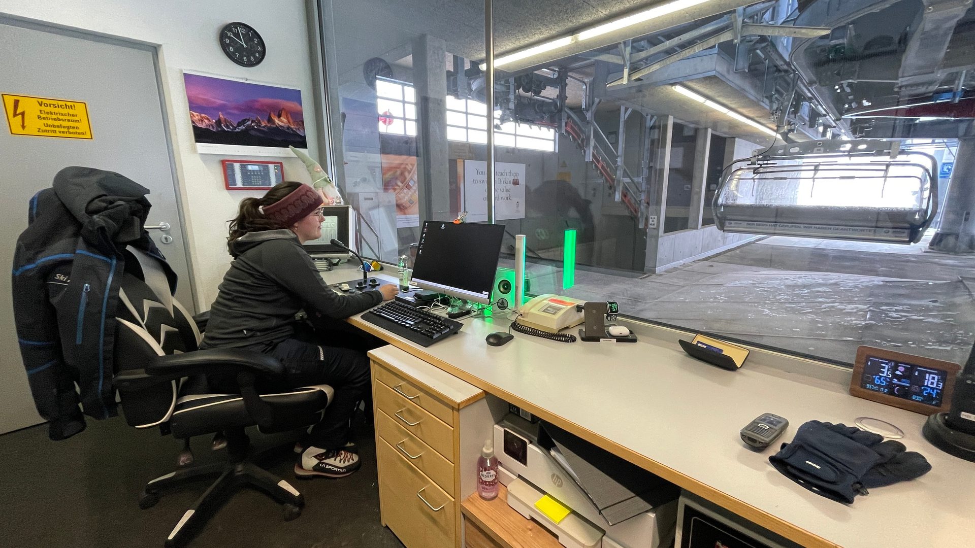 A ski lift operator works at a control desk, viewing the station and a chairlift through a window.