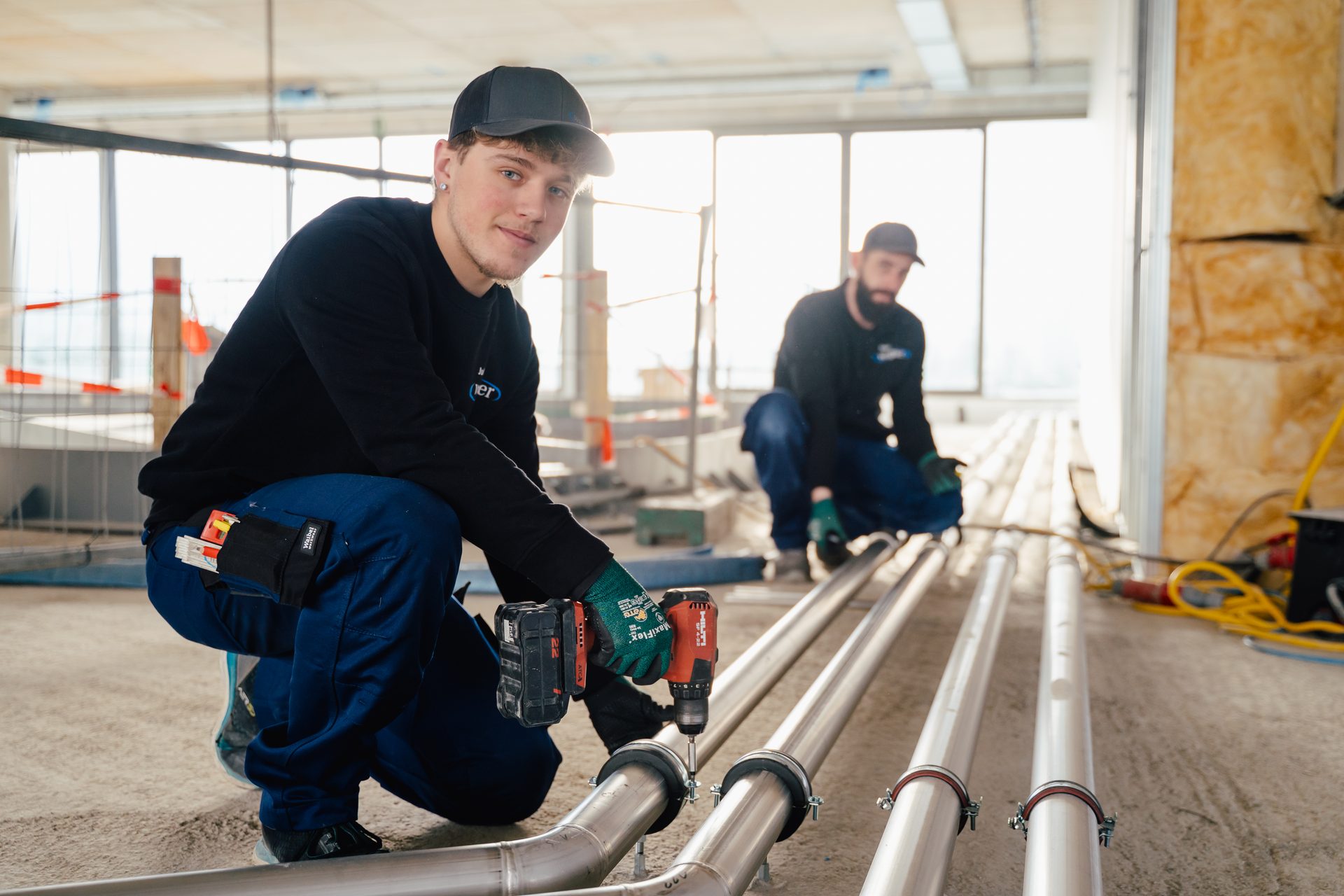 Two construction workers installing pipes on a site. One works with a drill, the other is in the background.