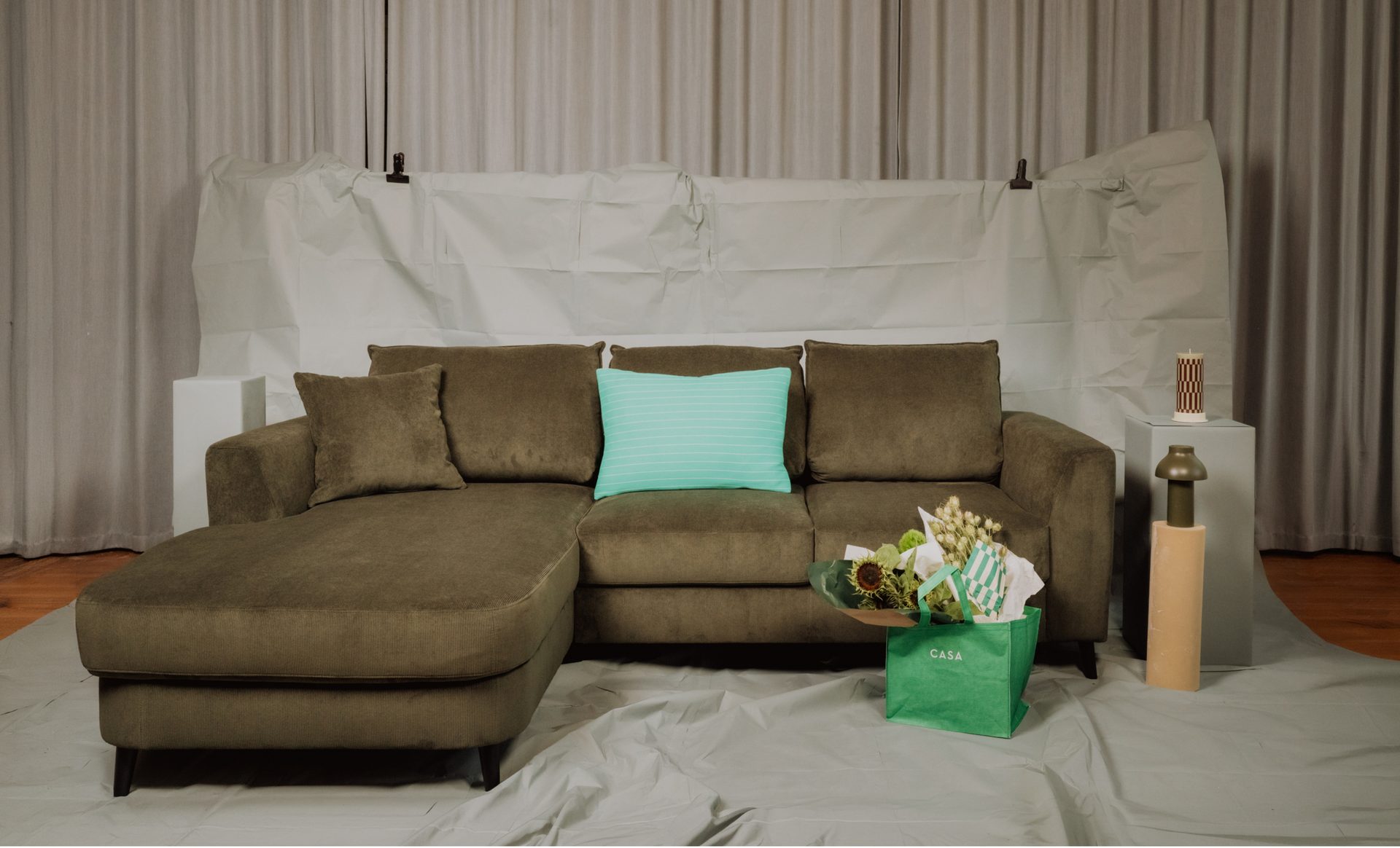 An L-shaped olive green couch with a turquoise pillow, a green gift bag, and decor on pedestals.