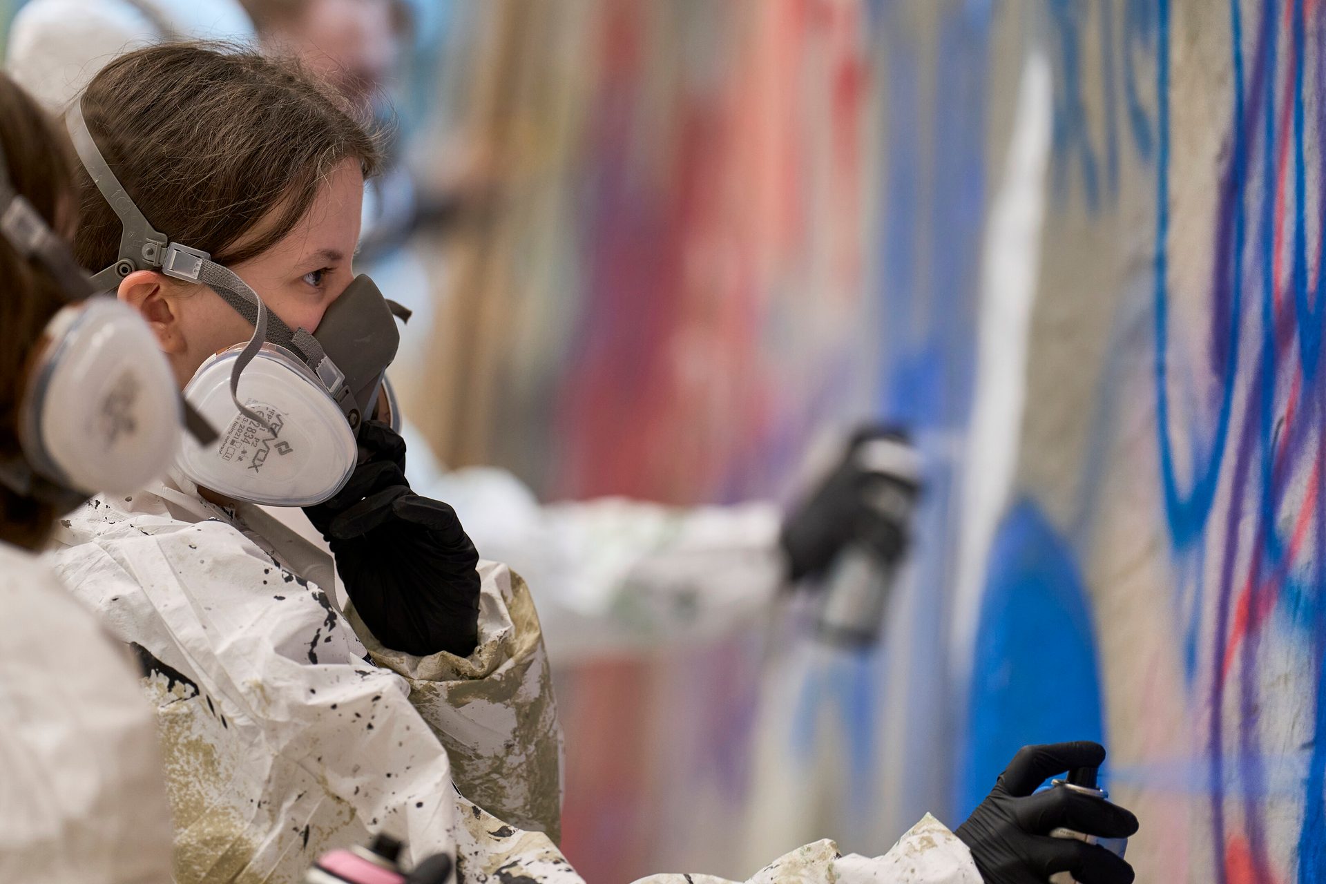 Two artists in protective gear spray painting a colorful graffiti mural, one in the foreground.