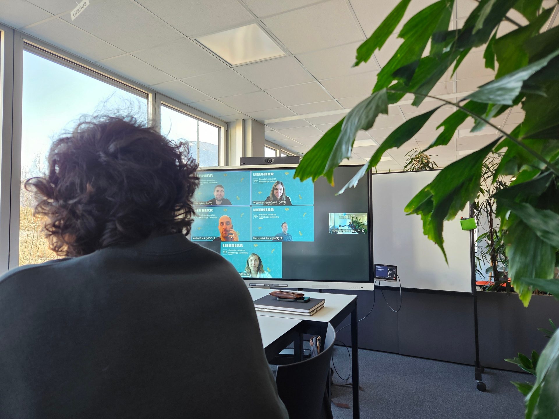 A person views a Liebherr video conference call displaying multiple participants in an office.
