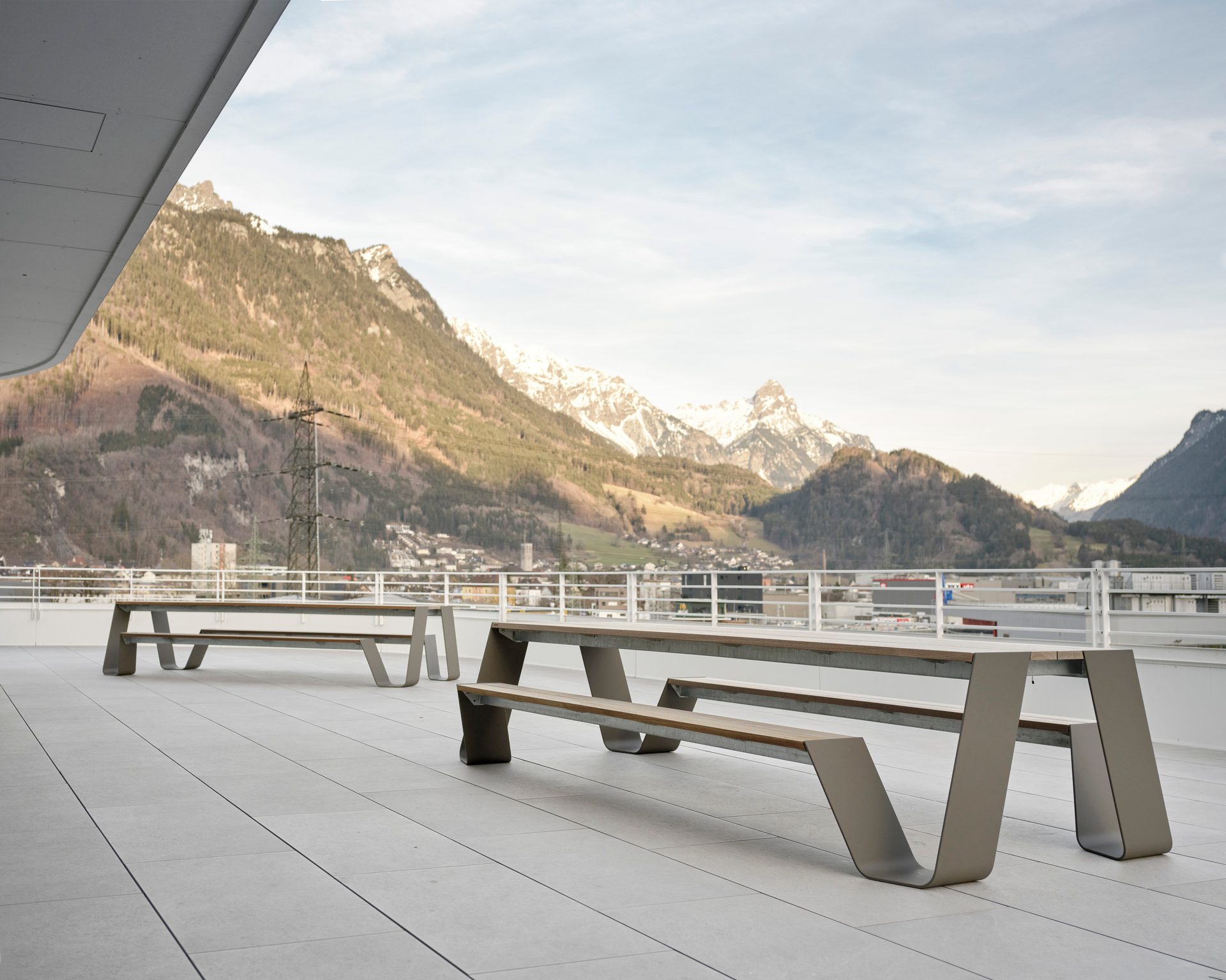 Modern outdoor picnic tables on a roof terrace with a stunning panoramic view of snow-capped mountains.
