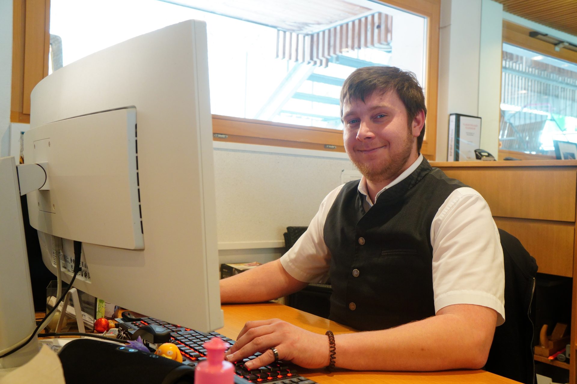 A smiling man in a black vest sits at a computer desk, hands on a keyboard.