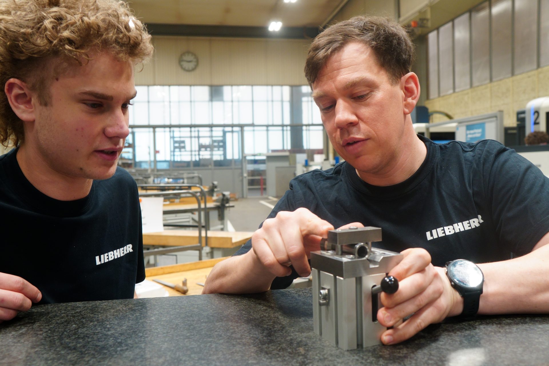 Two men wearing black 'Liebherr' shirts examining a piece of machinery on a speckled table.