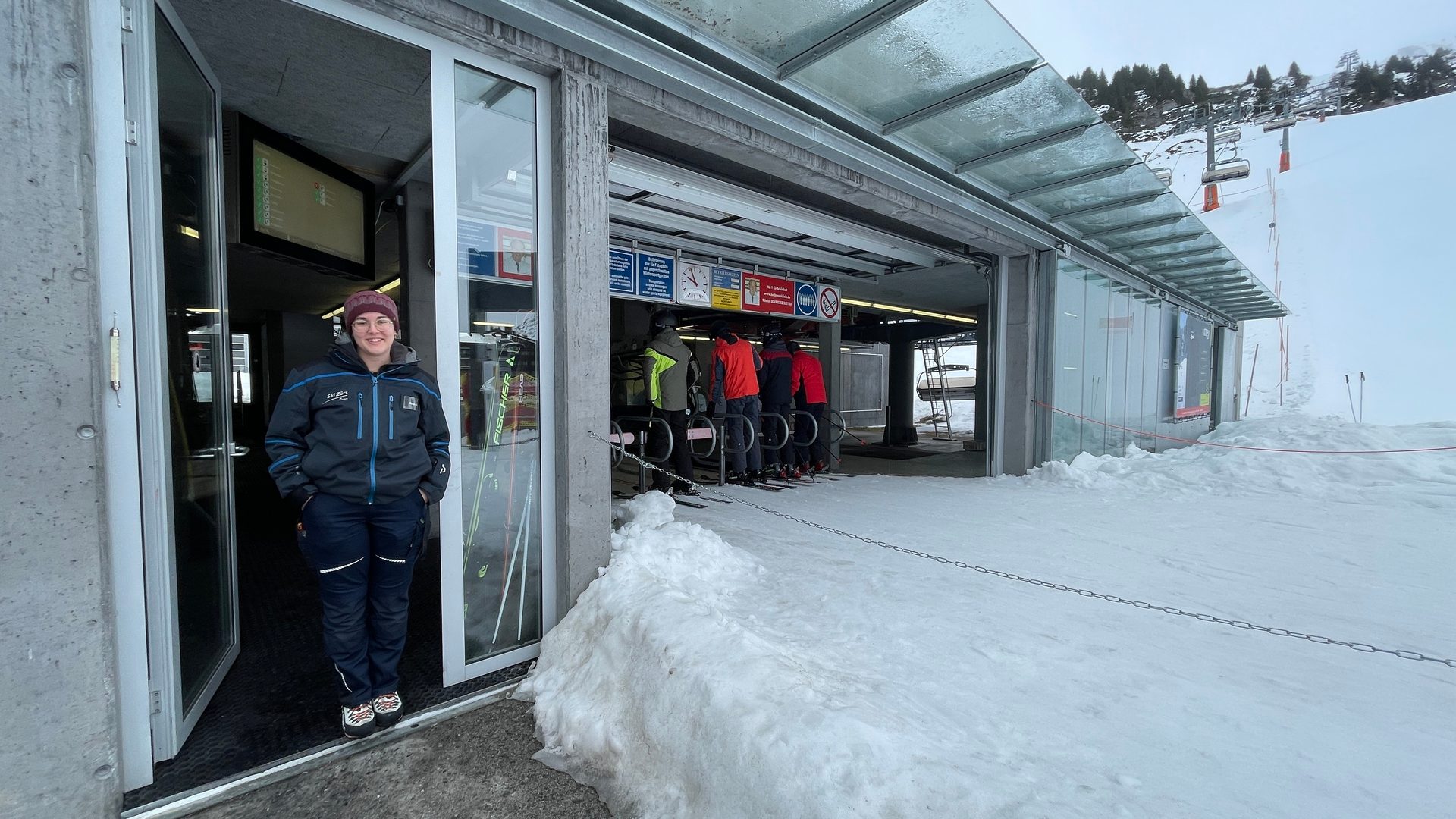Woman in uniform at ski lift entrance; skiers, snow, chairlifts in background.