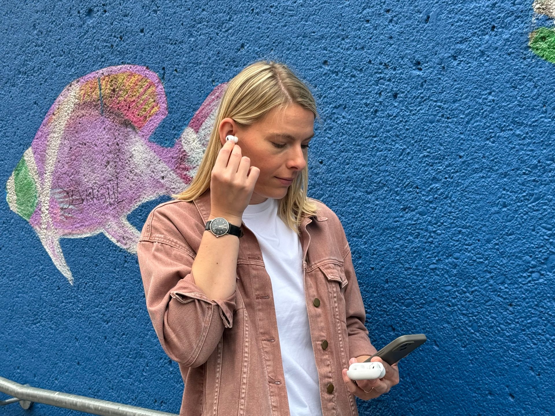 Woman putting in earbud, holding phone and case, blue mural background.
