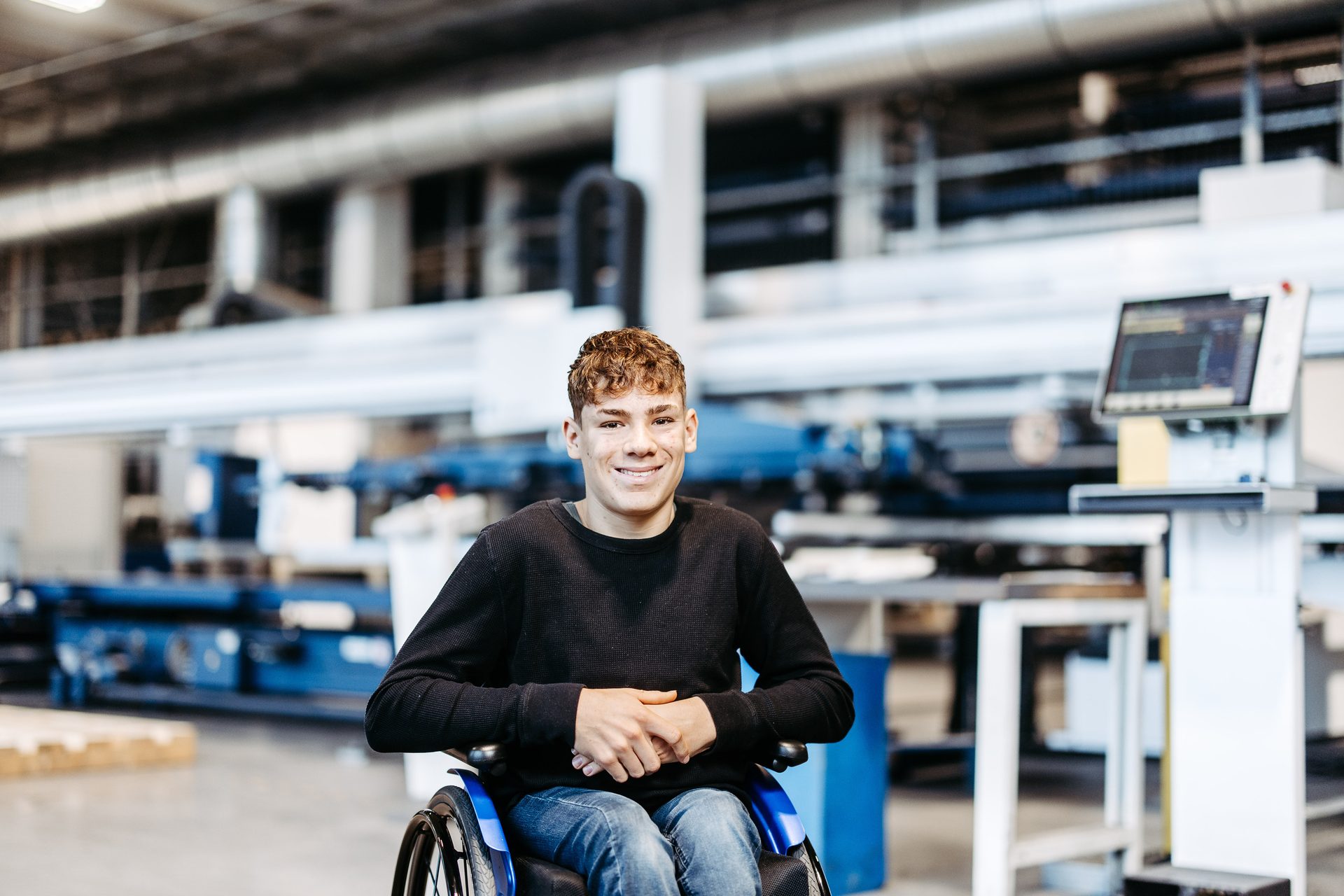 A smiling young man with curly hair in a wheelchair, seated in a modern factory or workshop.