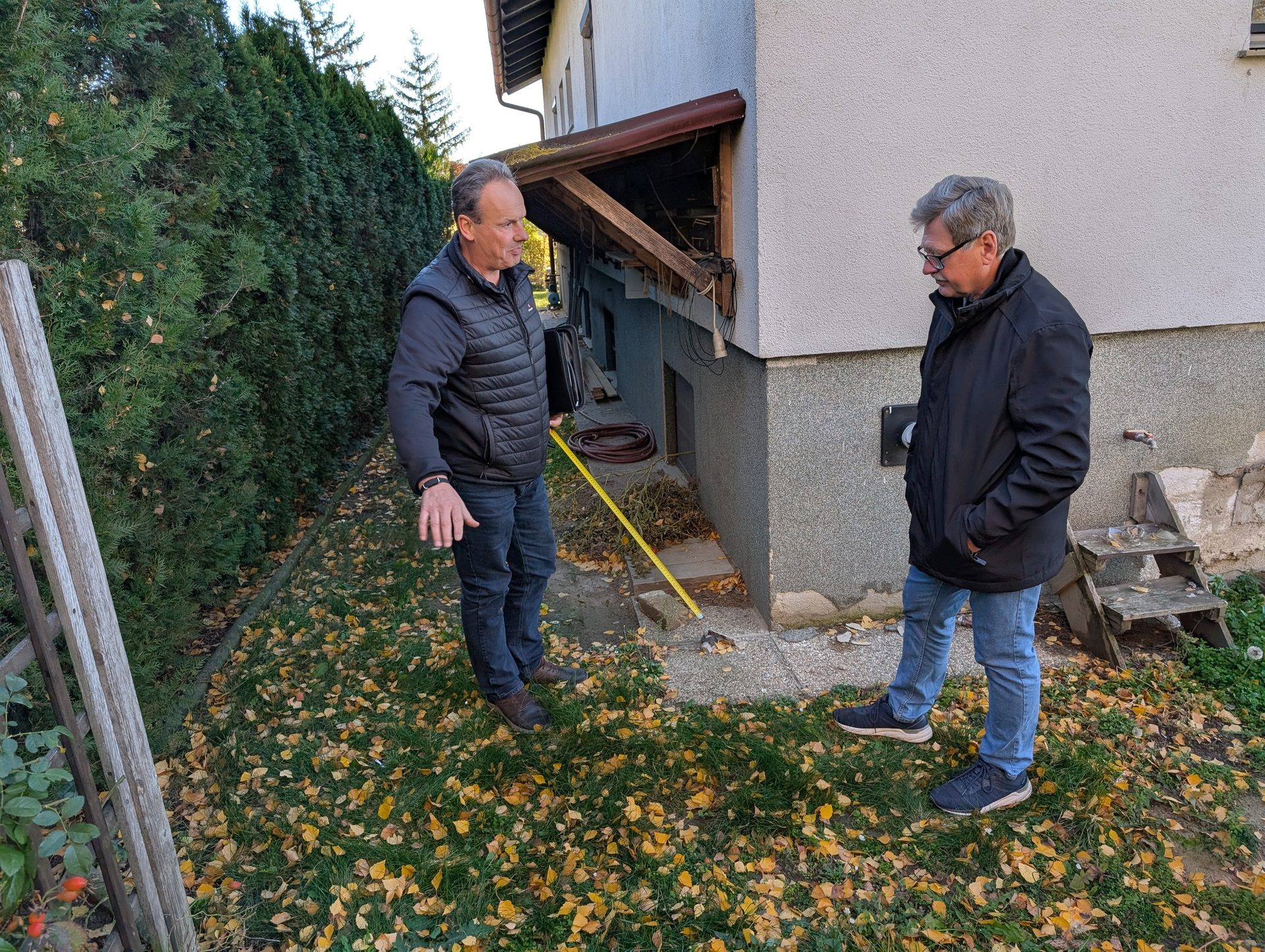Two men with a measuring tape beside a house, surrounded by autumn leaves.