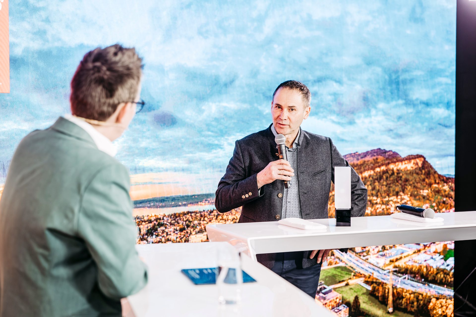 Two men at a presentation; one speaks into a microphone, the other listens. Scenic screen background.