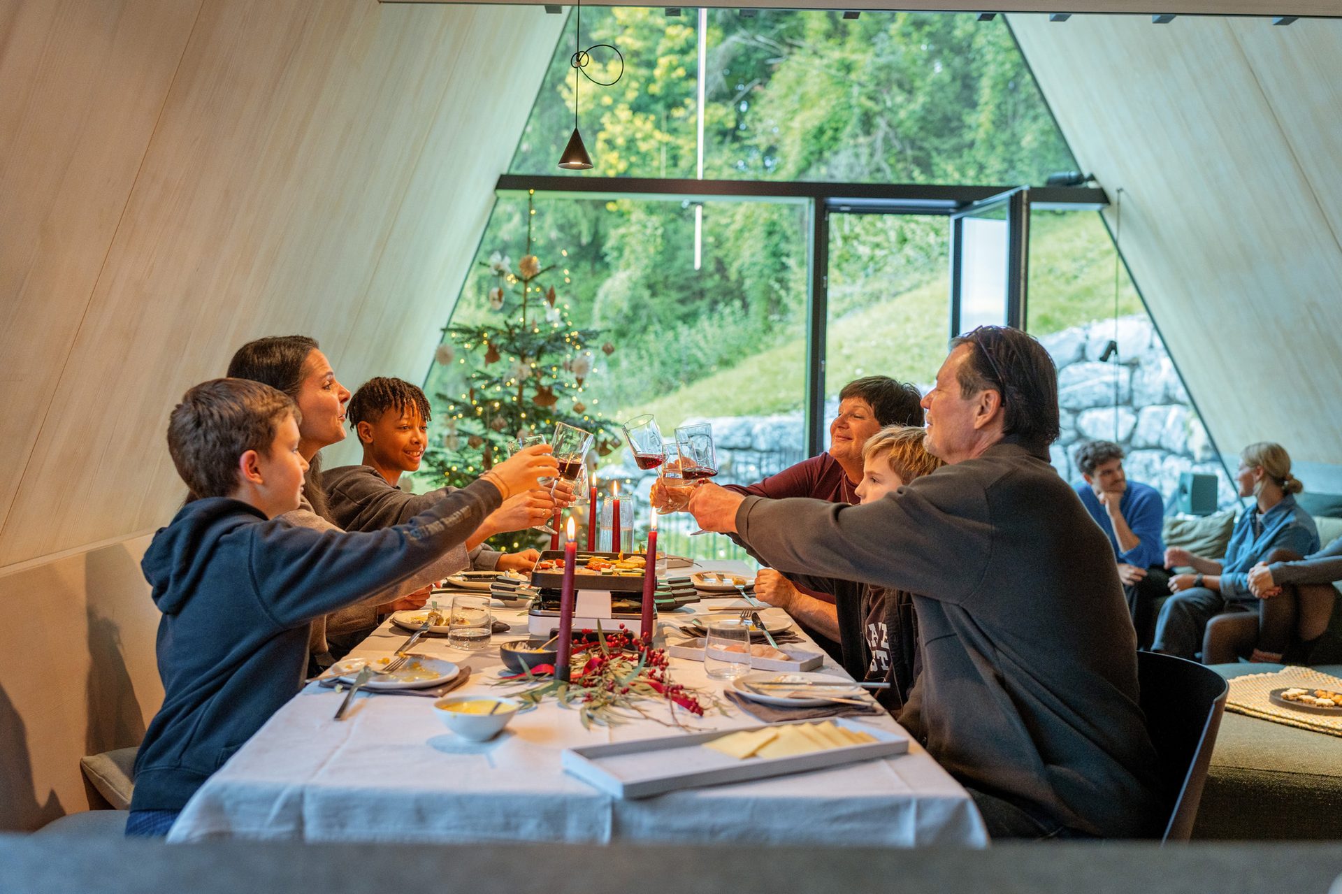 Family toasting at a festive dinner table in a modern house with a Christmas tree and mountain view.