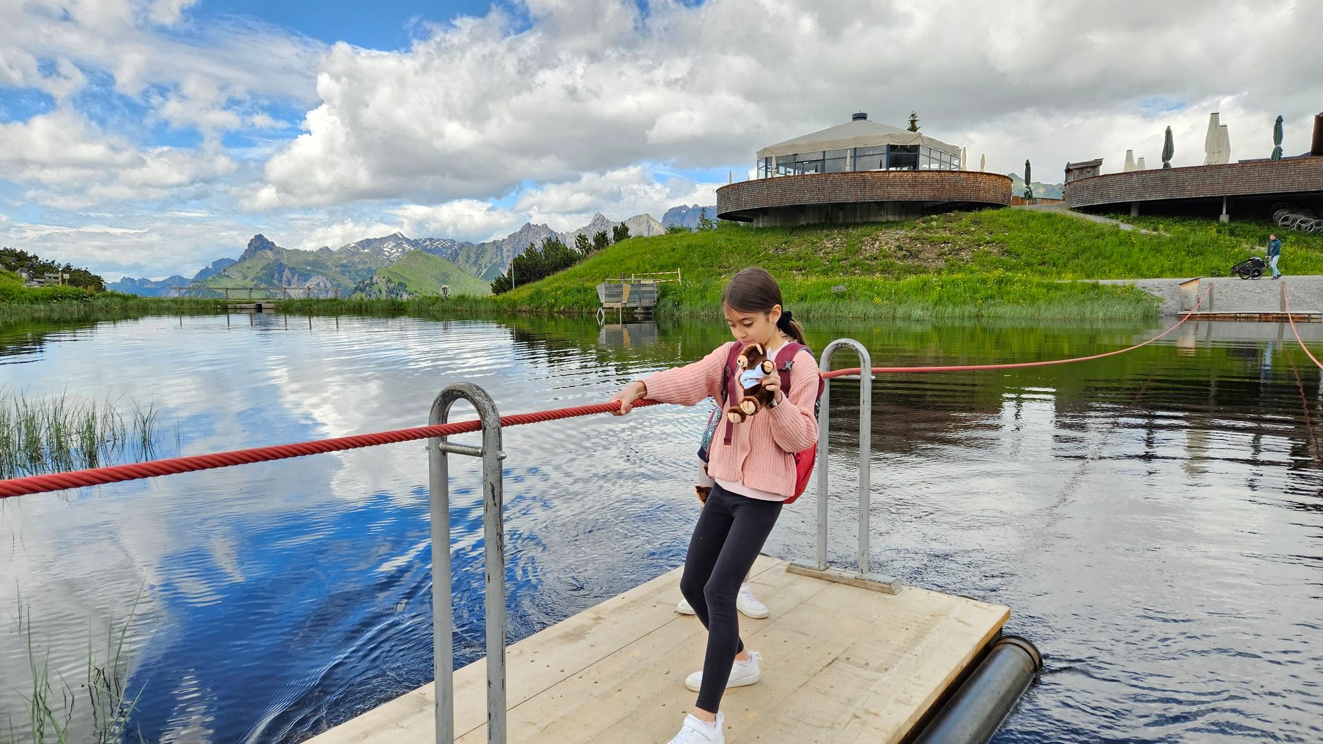 A girl holds a toy on a lake platform, with mountains and a modern building in the background under cloudy skies.