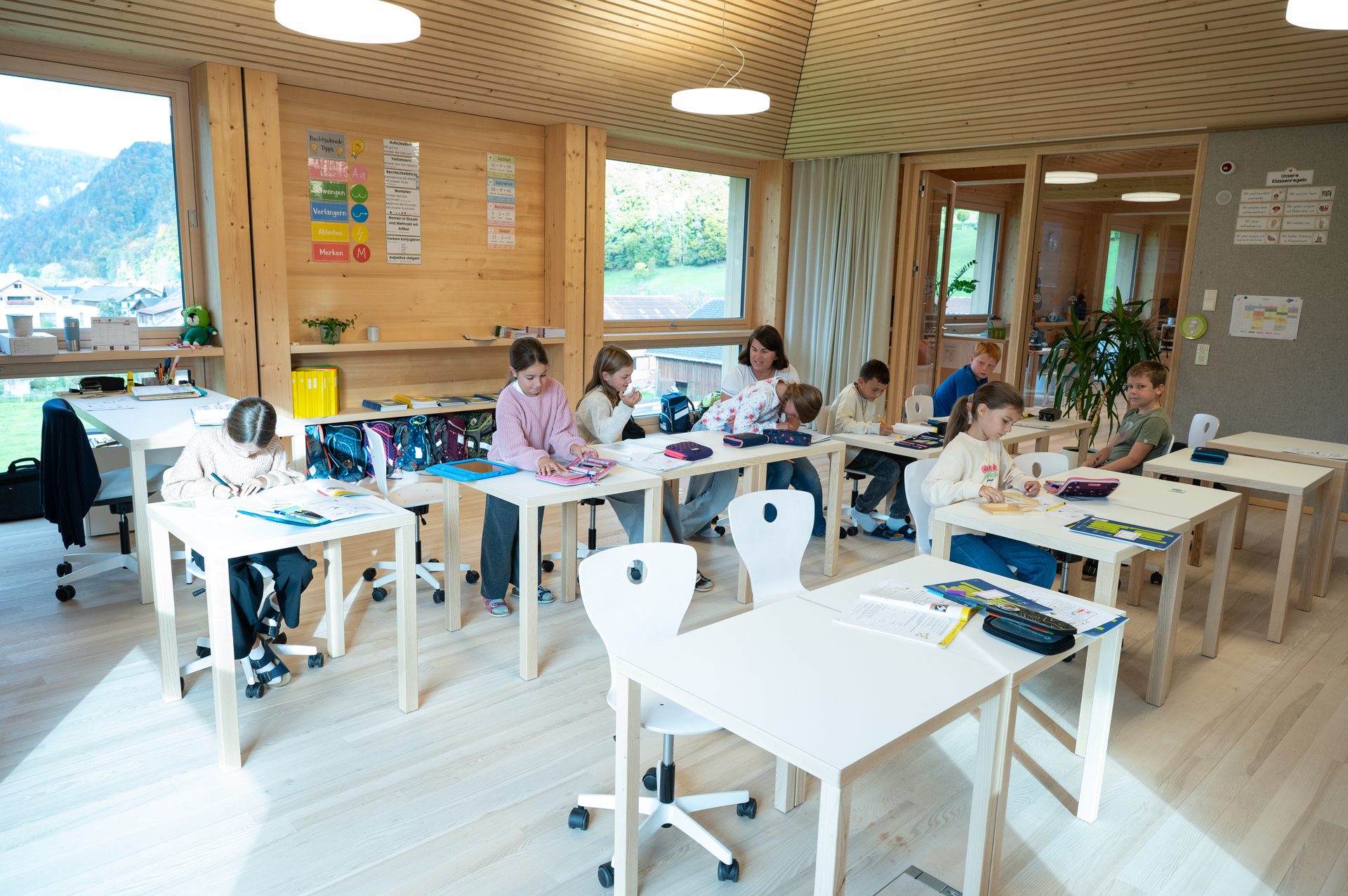 Primary school children and a teacher in a bright, modern wooden classroom, working at desks.