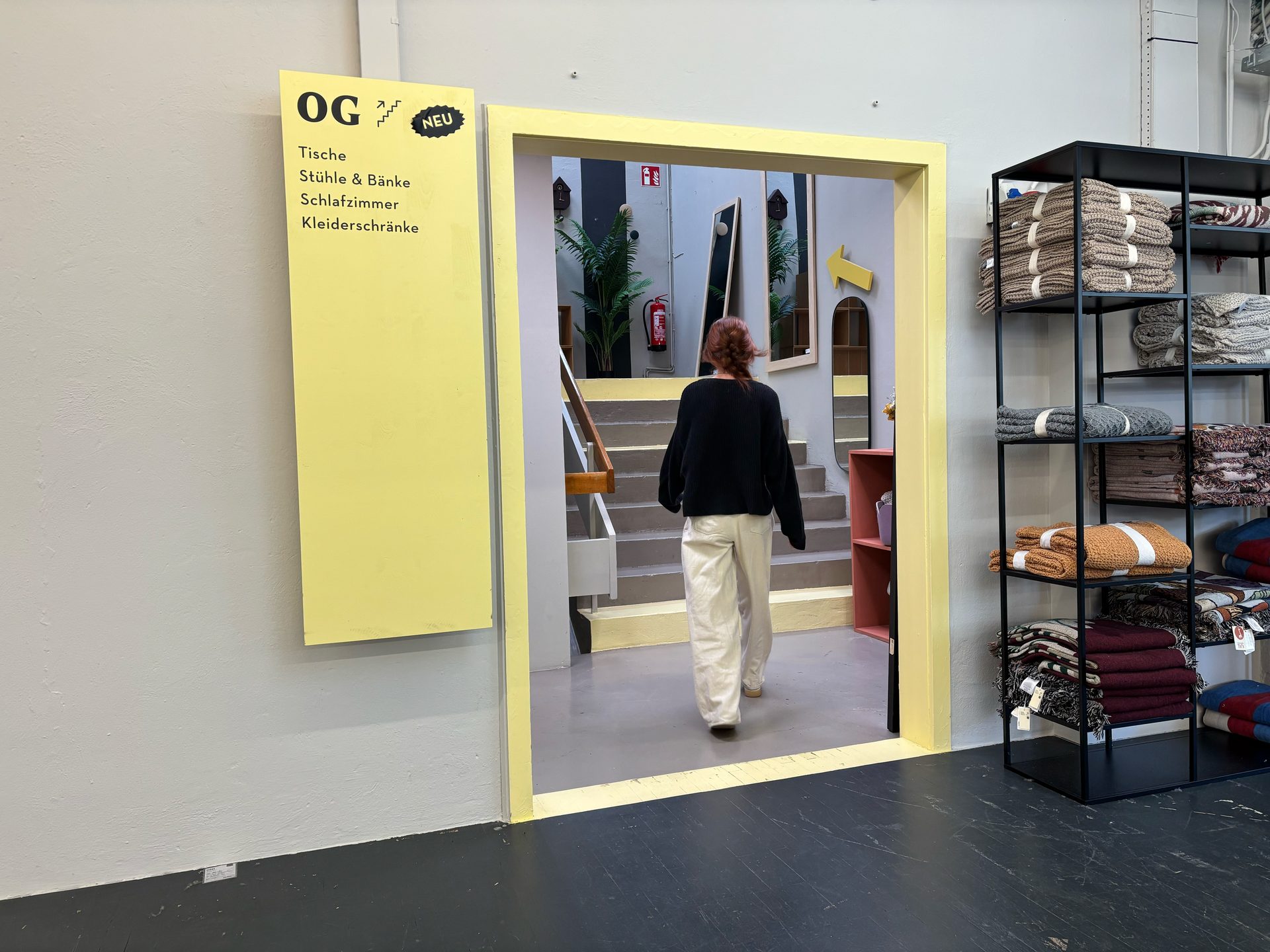 A person walks through a yellow doorway next to a yellow sign listing furniture departments and a shelf of textiles.