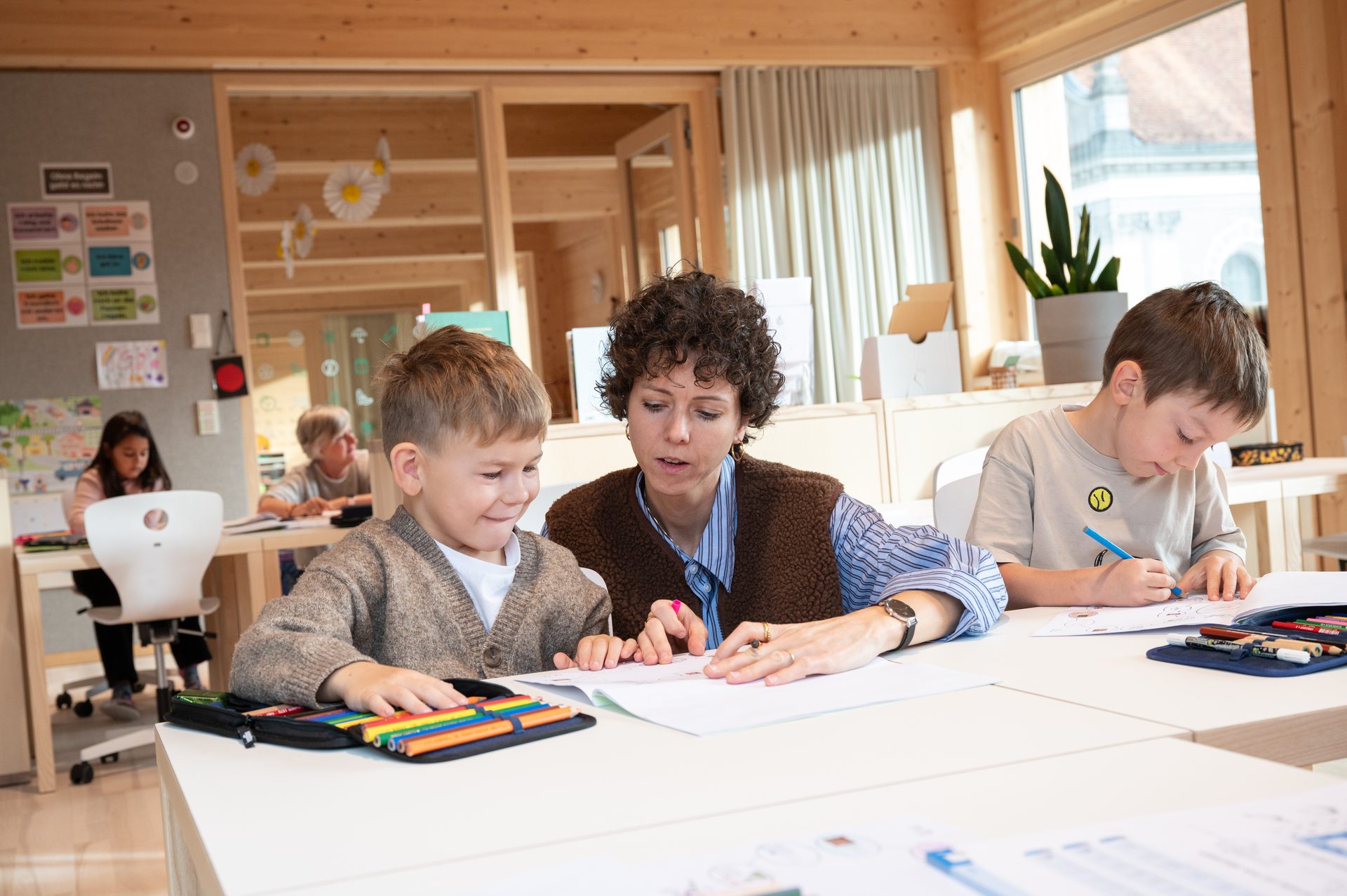 Teacher helps a boy with schoolwork, another boy draws in a bright classroom.