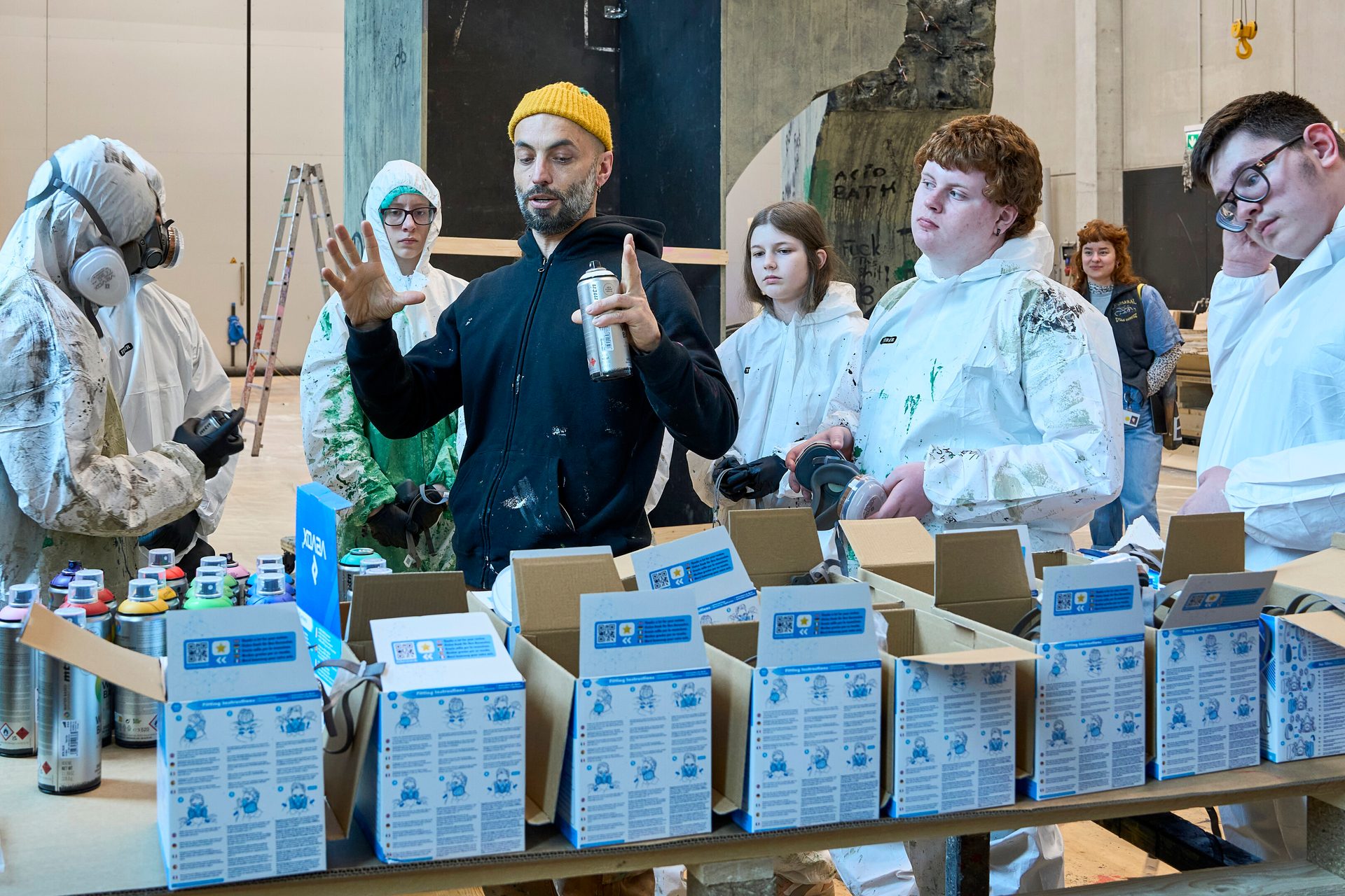 Instructor in yellow hat with spray can teaches students in protective suits, surrounded by workshop supplies.