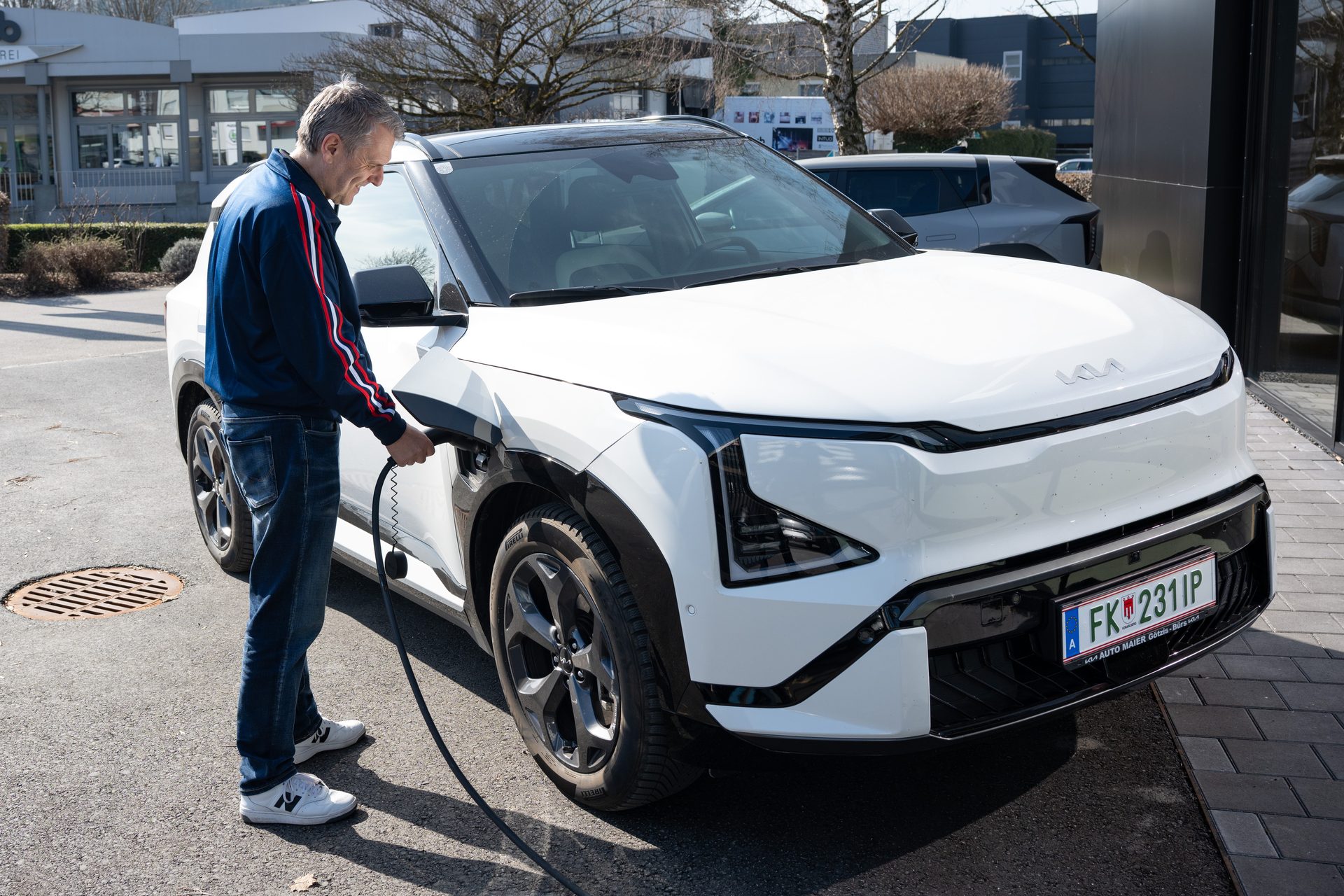 Man charging a white Kia EV9 electric SUV outdoors during the day.