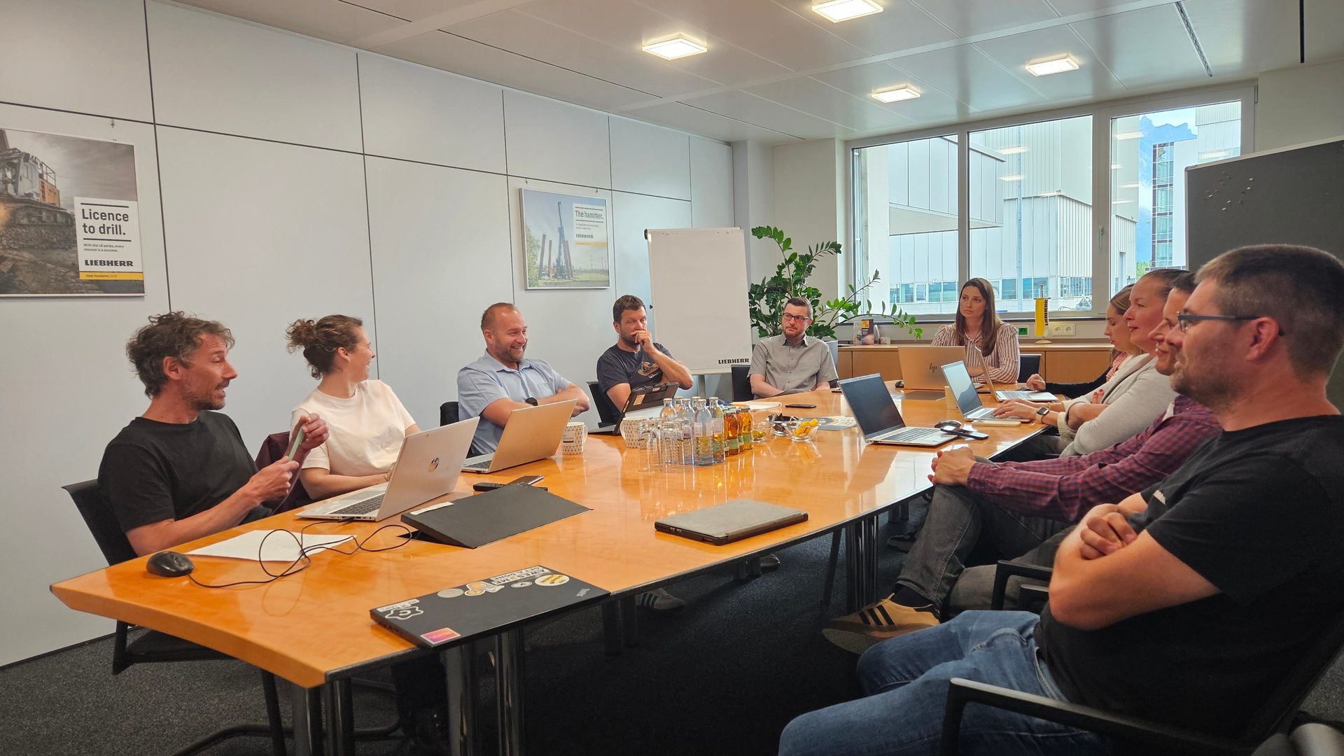 Diverse team collaborating at a large wooden table with laptops and drinks in a bright meeting room.