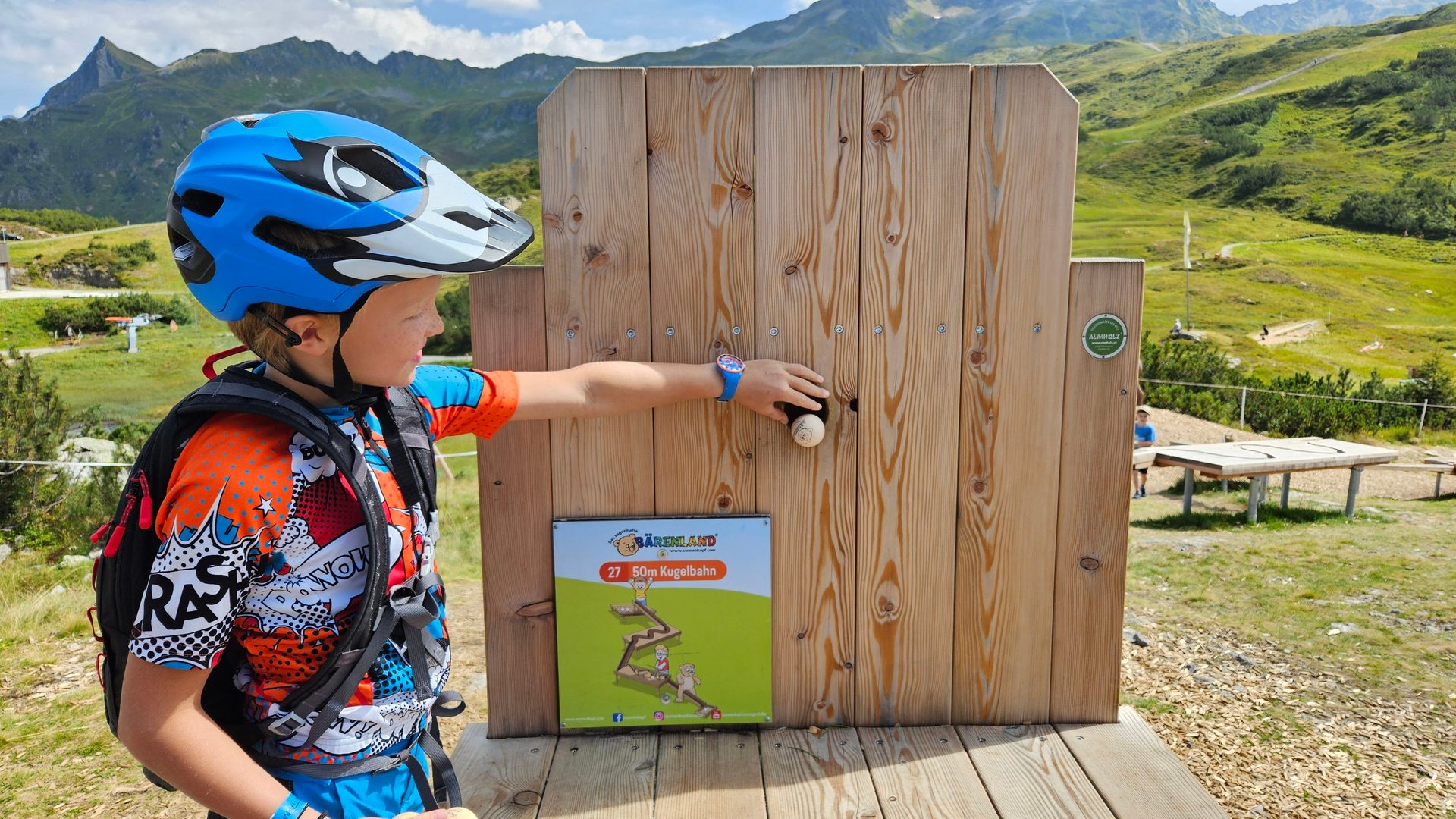 A child in a bike helmet and backpack plays a wooden ball track game in a scenic mountain setting.