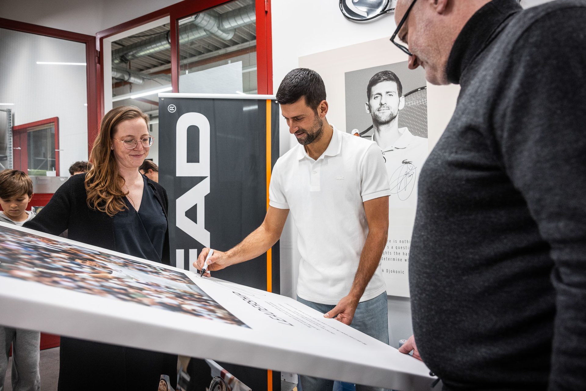 Novak Djokovic signs an item as a woman and another man observe. A "HEAD" banner is in the background.