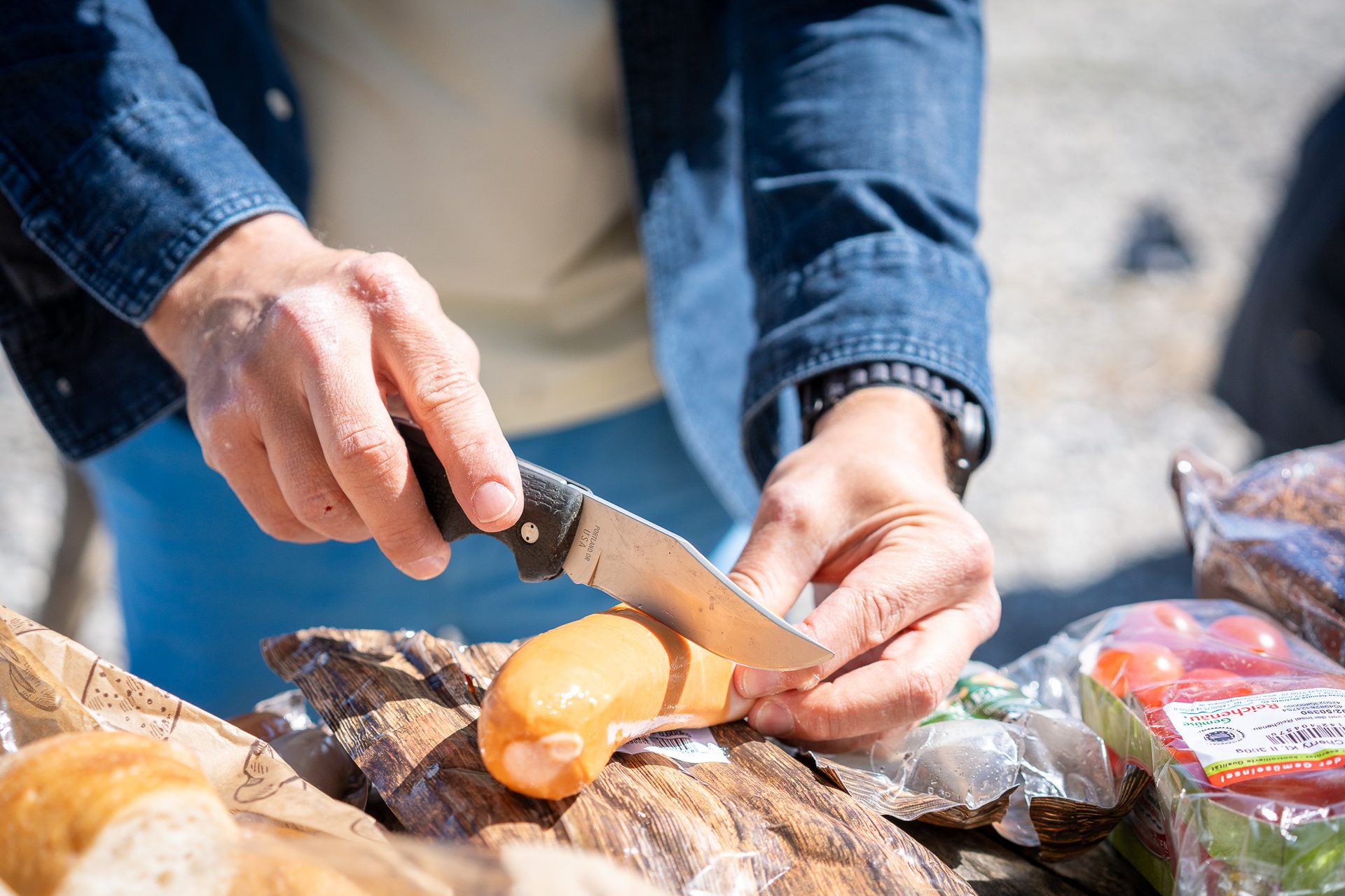 A person uses a knife to cut a sausage on a wooden surface, with other picnic items nearby.