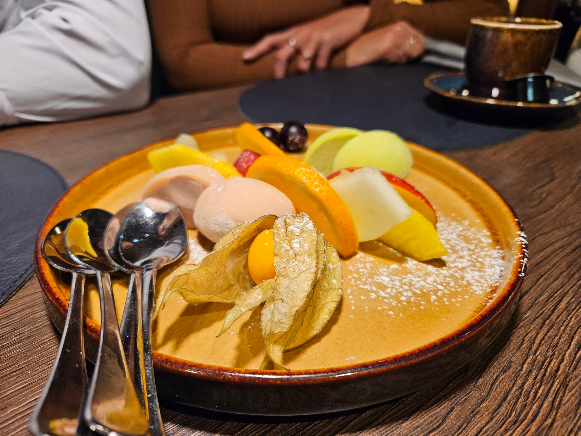 Dessert plate with fruits, mochi, and spoons, with a person and coffee cup in background.