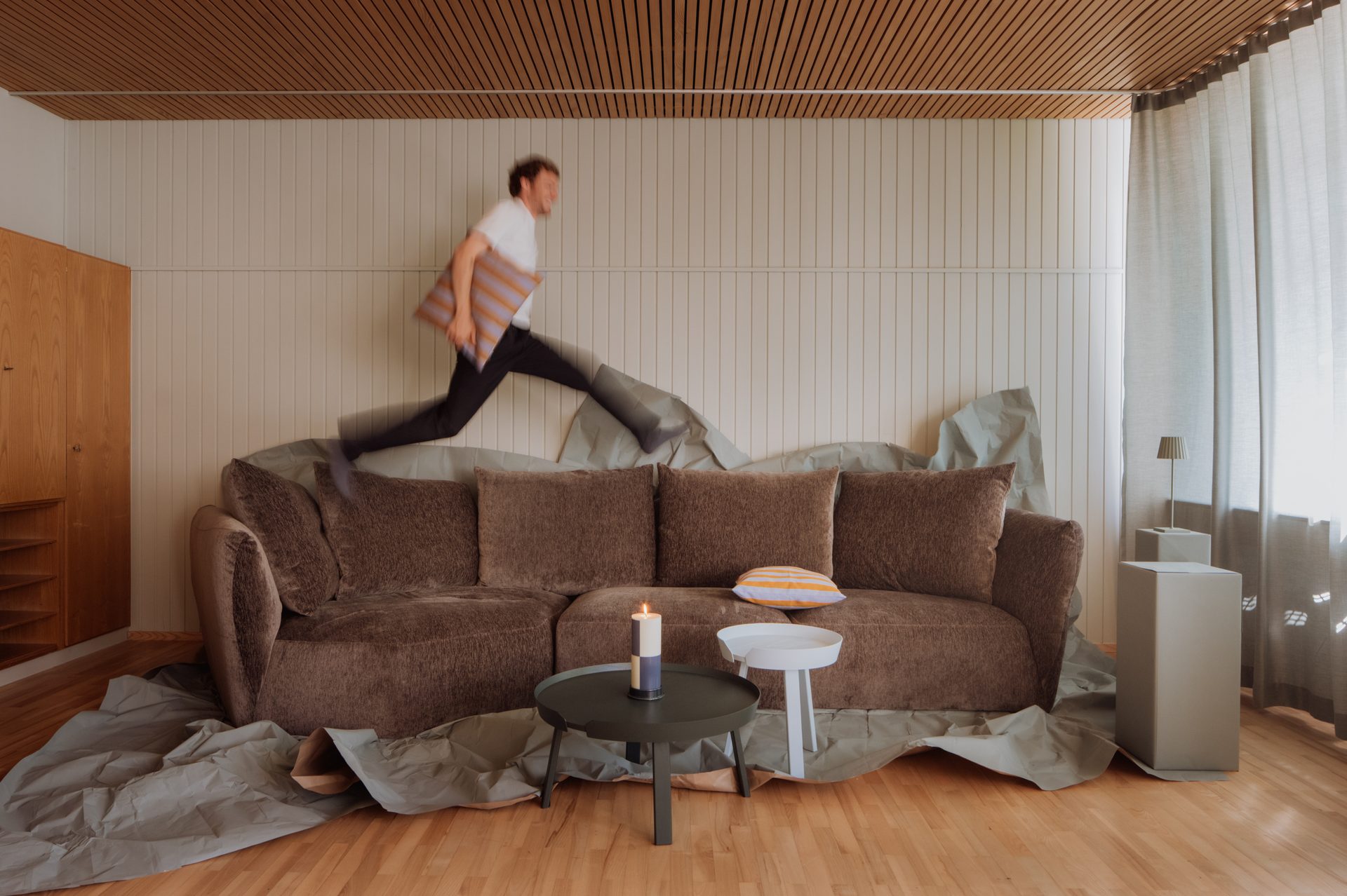 A man jumps over a brown sofa, holding a striped pillow, with side tables and a candle in a modern room.