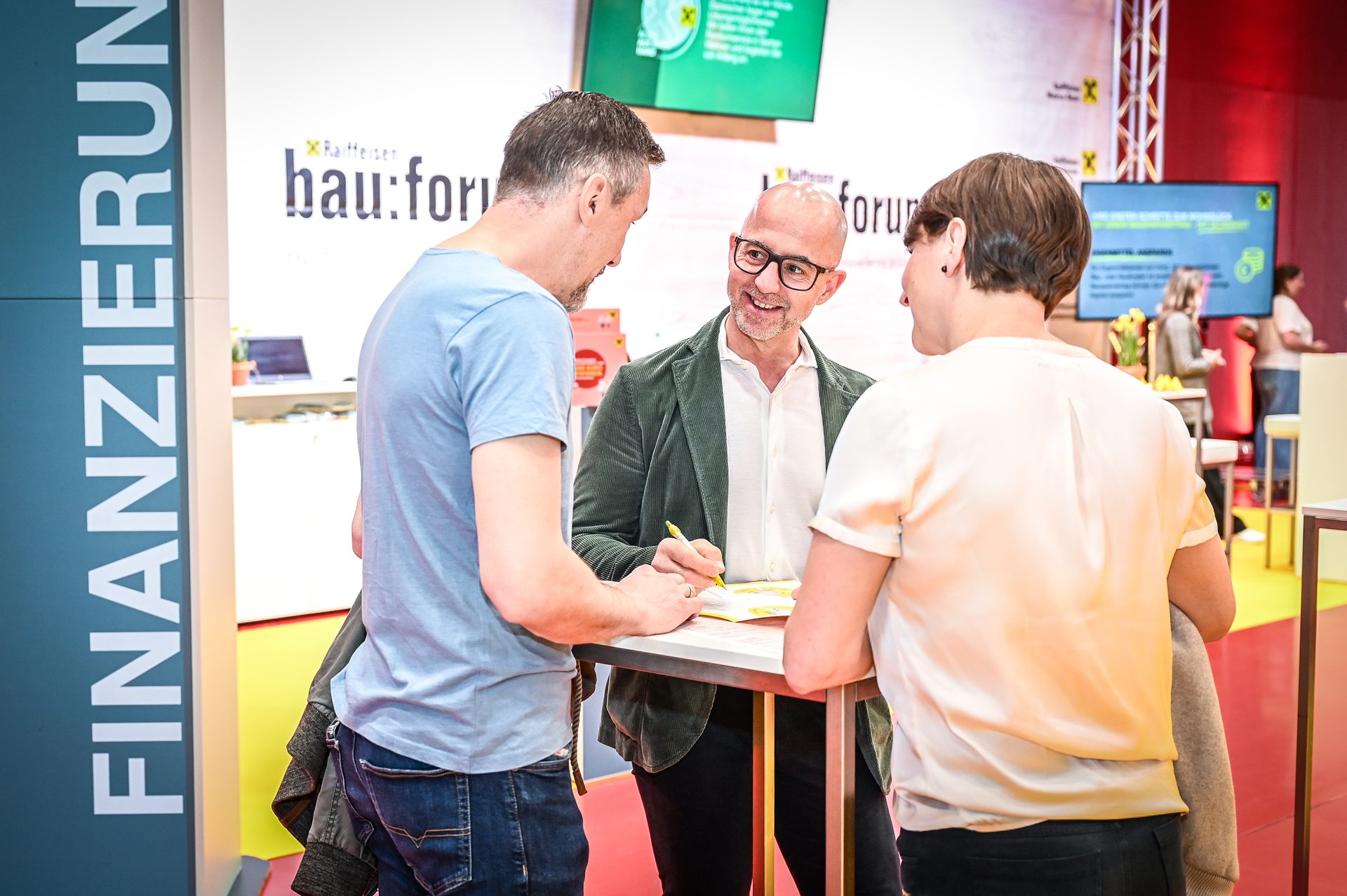 Three people, two men and one woman, are conversing and writing at a business event booth.