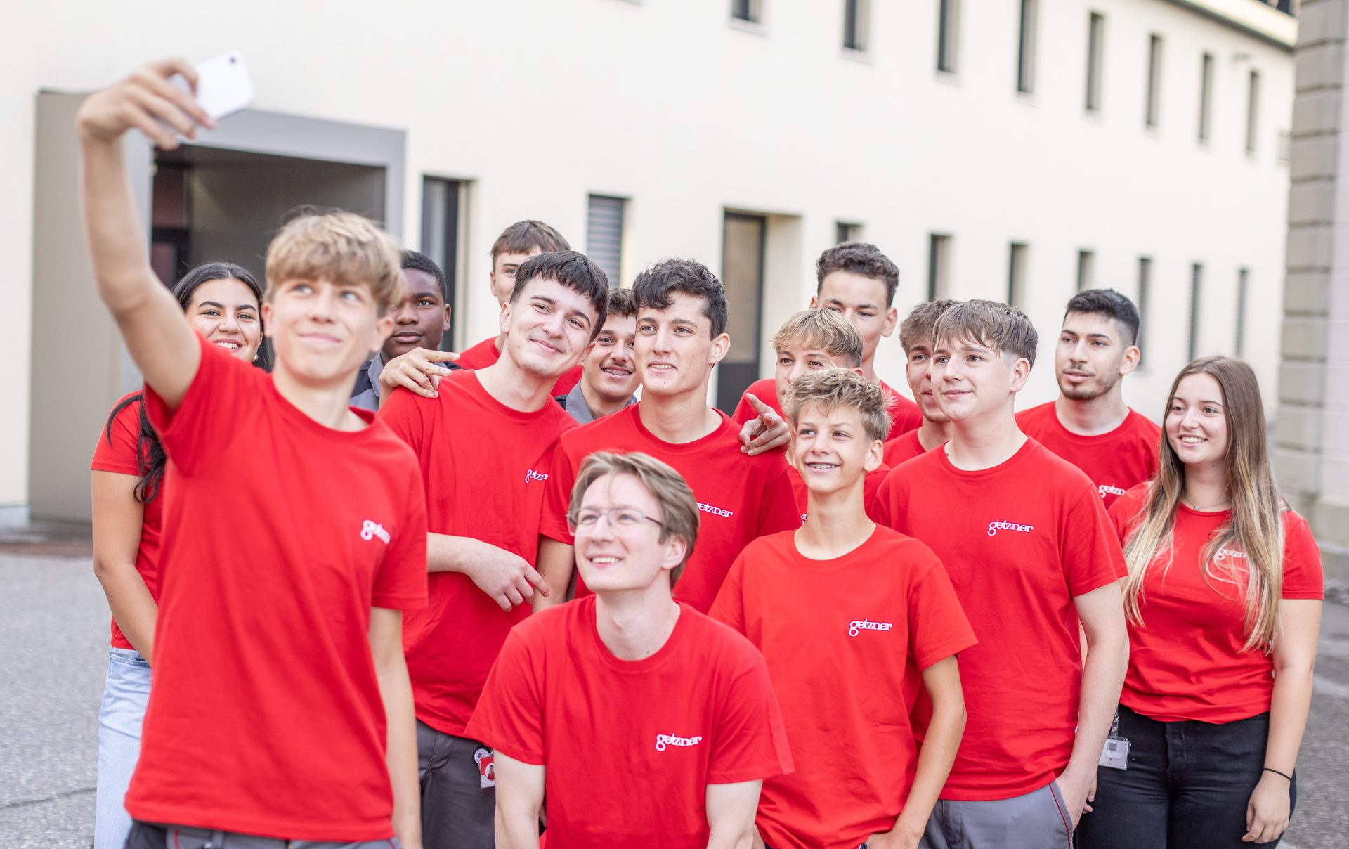 A diverse group of young people in red "getzner" shirts takes a selfie outdoors.