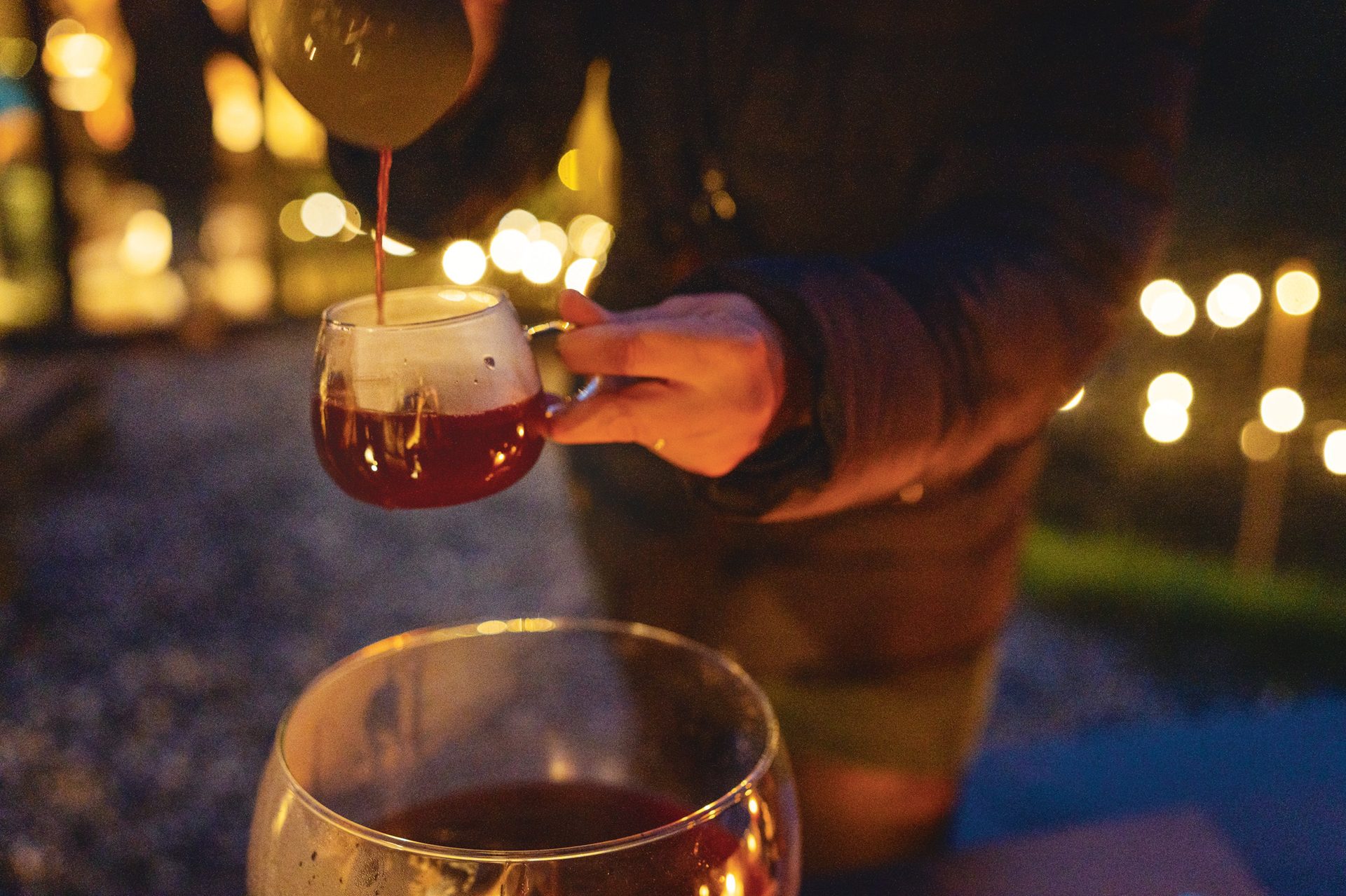 A hand pours a red drink from a jug into a glass mug, with blurred golden lights in the background.