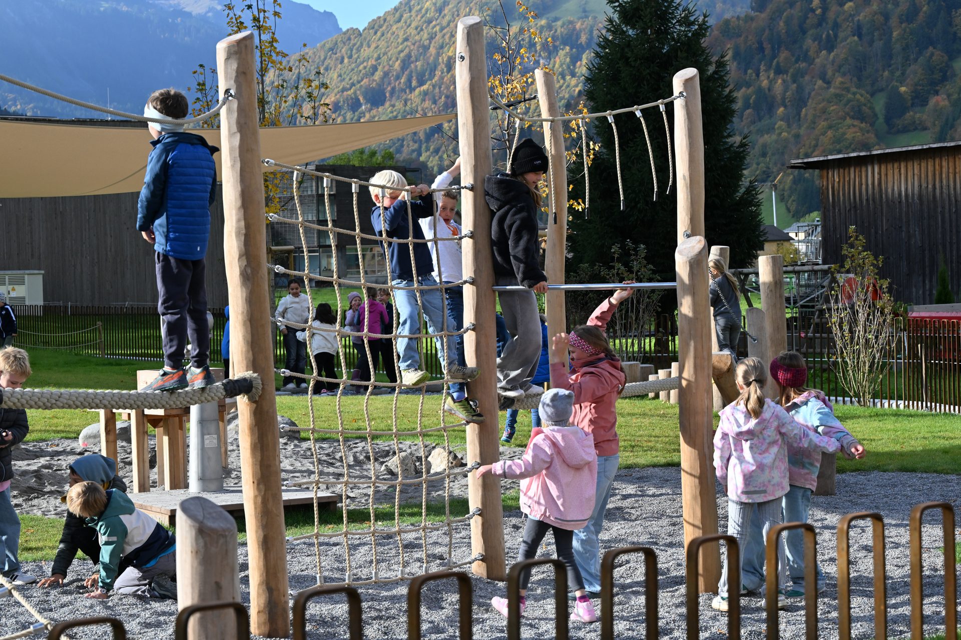 Children play on a wooden and rope climbing structure at a playground with mountains behind.