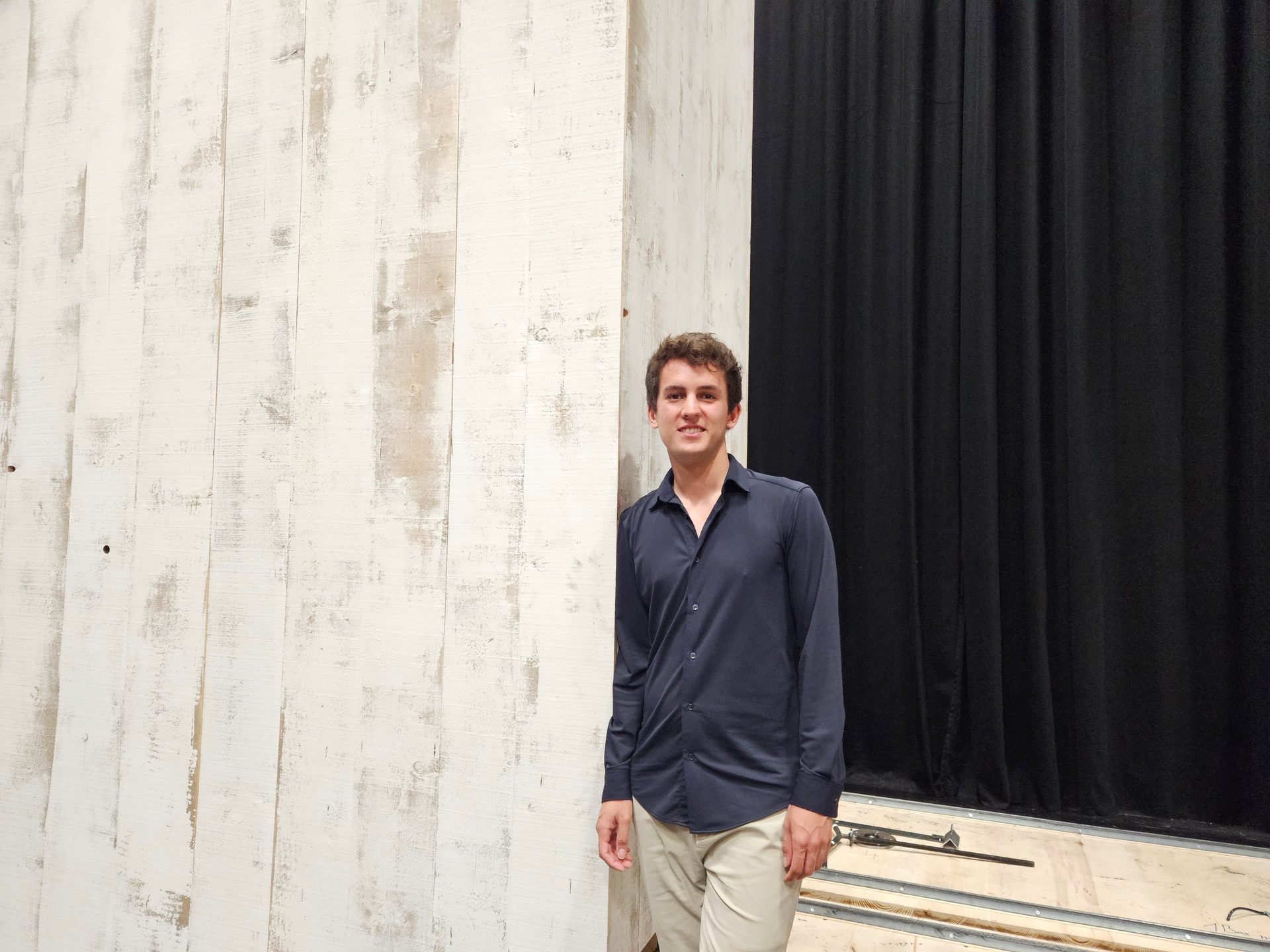A young man smiles, standing beside a white wooden wall with a black curtain behind him.