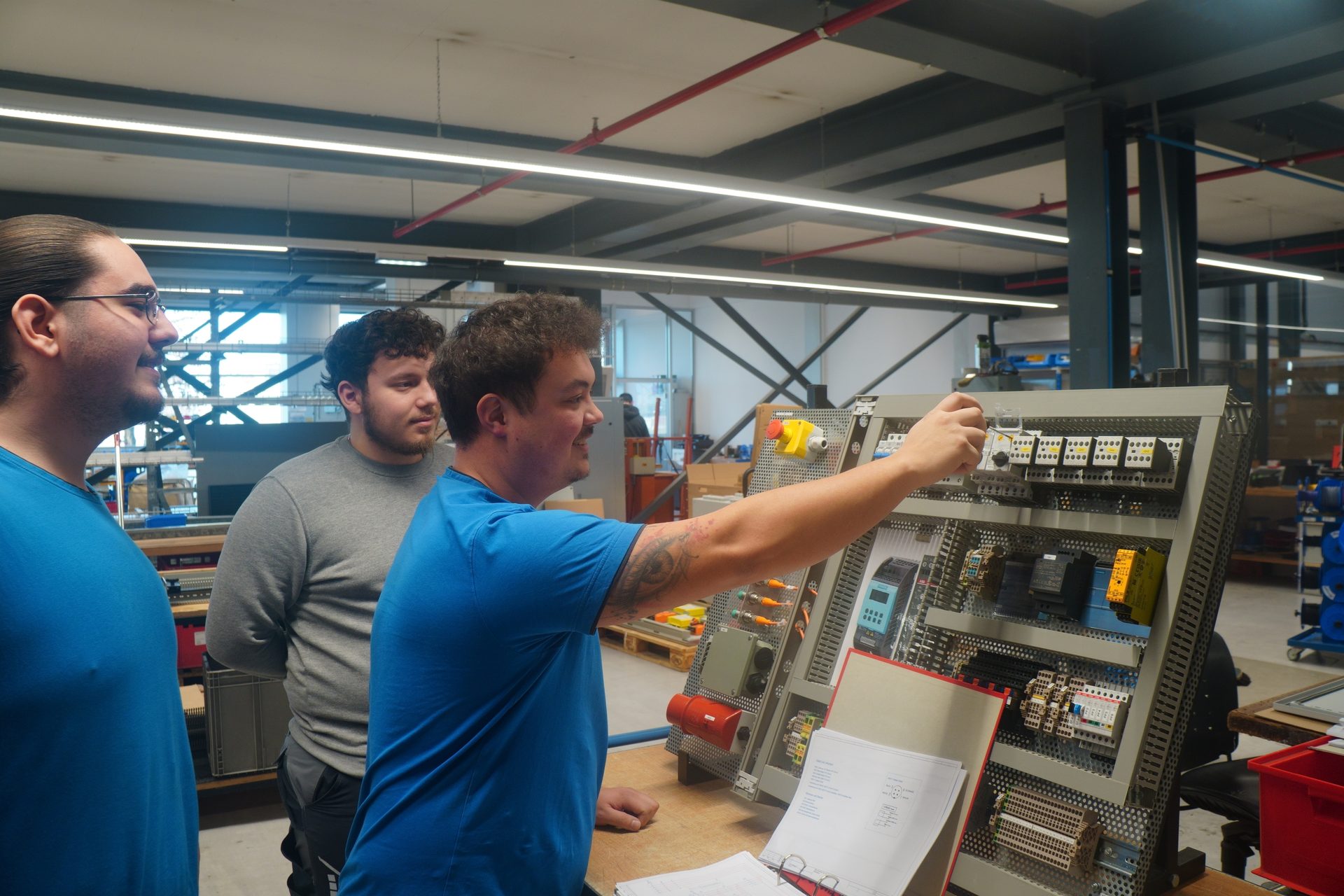Three men in a workshop, one demonstrating or learning with an electrical control panel.