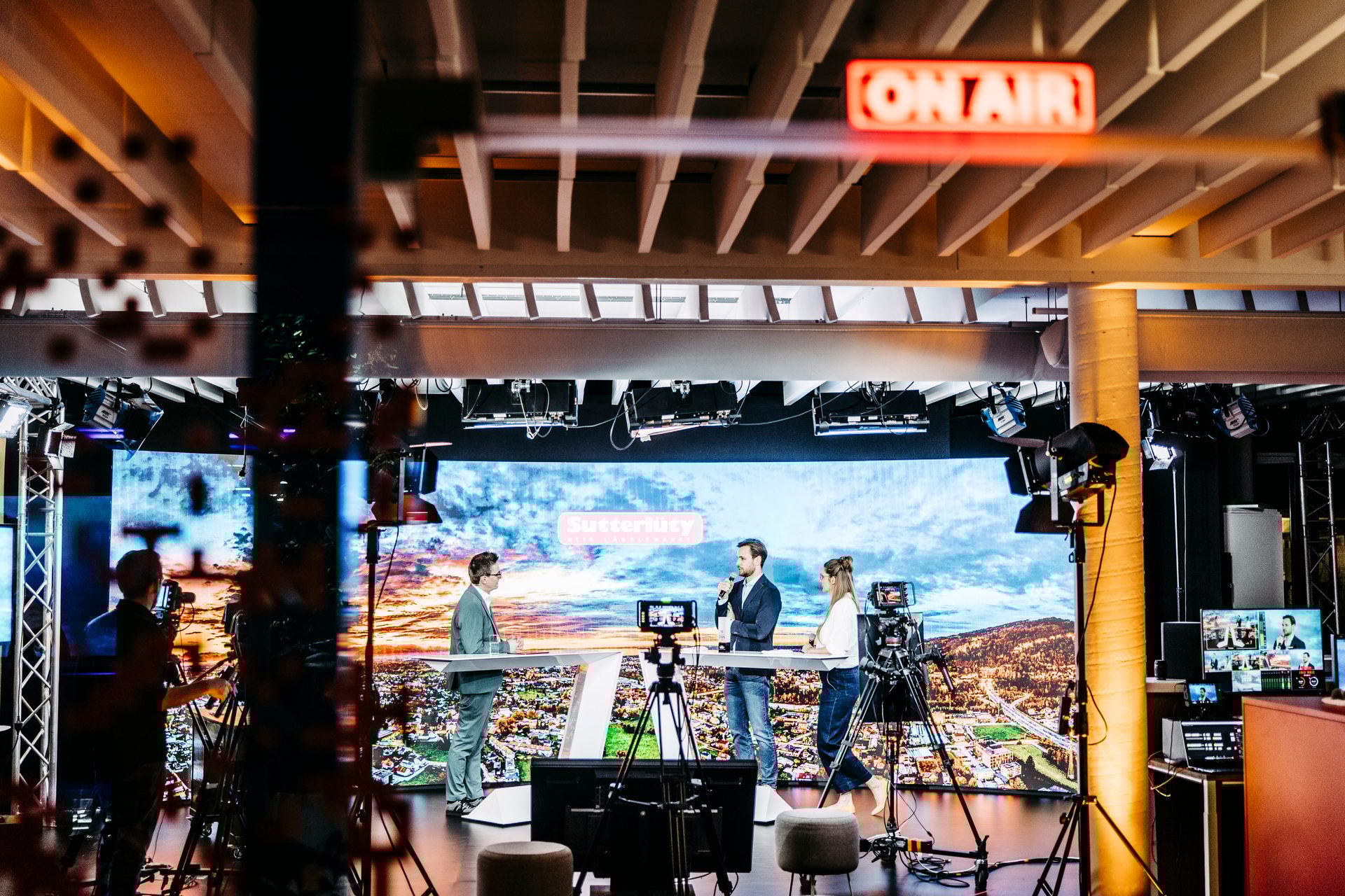 Three people broadcast live in a TV studio, with an "ON AIR" sign and a large digital backdrop.