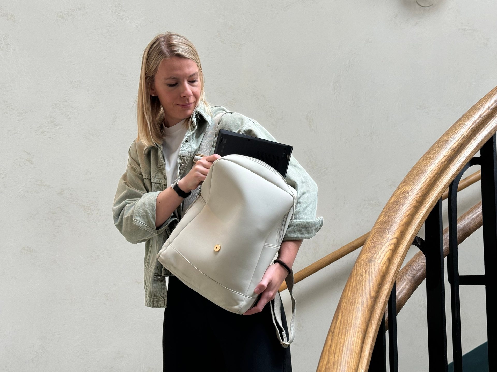 Woman puts laptop into light backpack near wooden staircase.