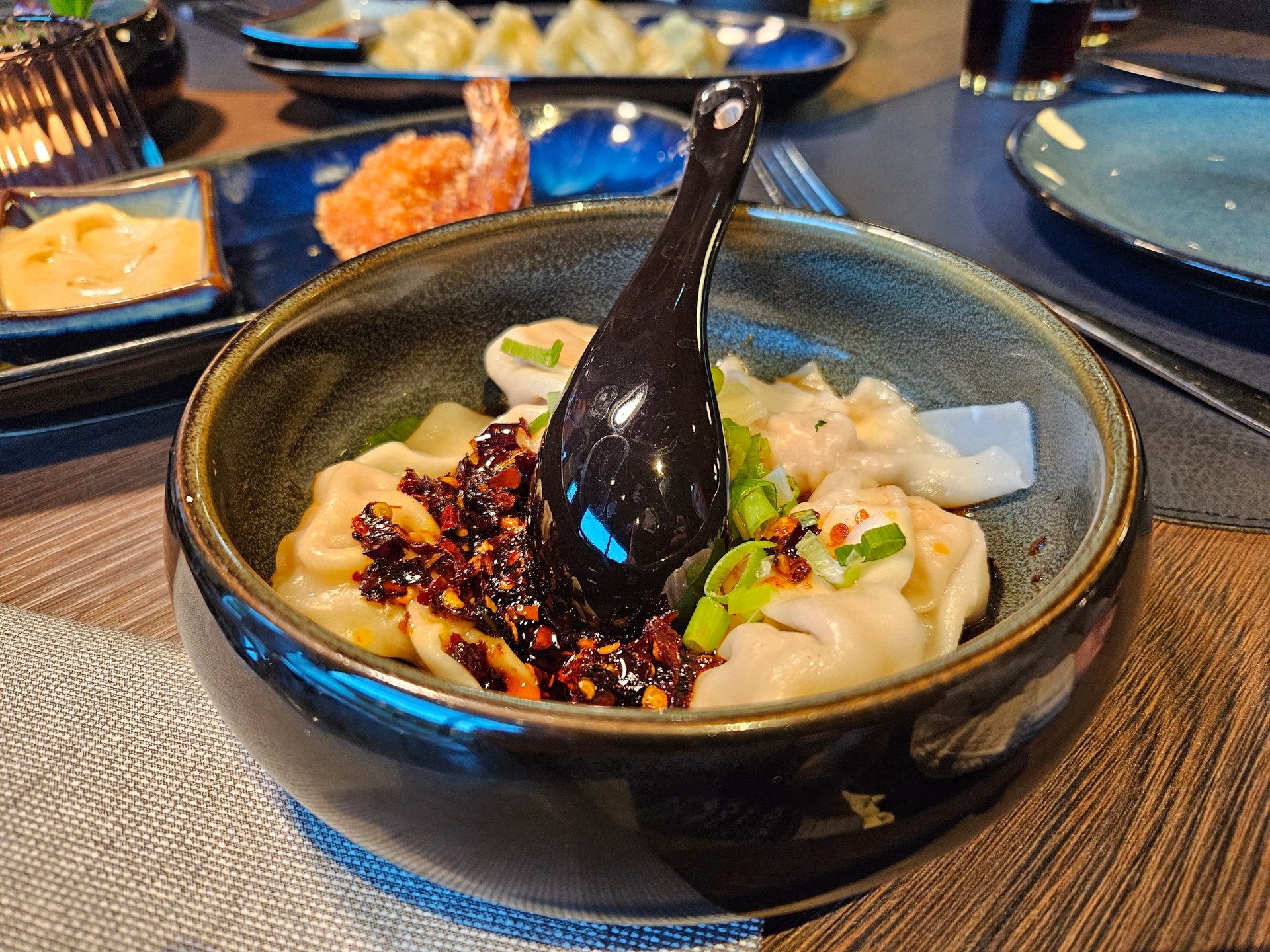 A dark bowl filled with dumplings topped with chili oil, green onions, and a black spoon.