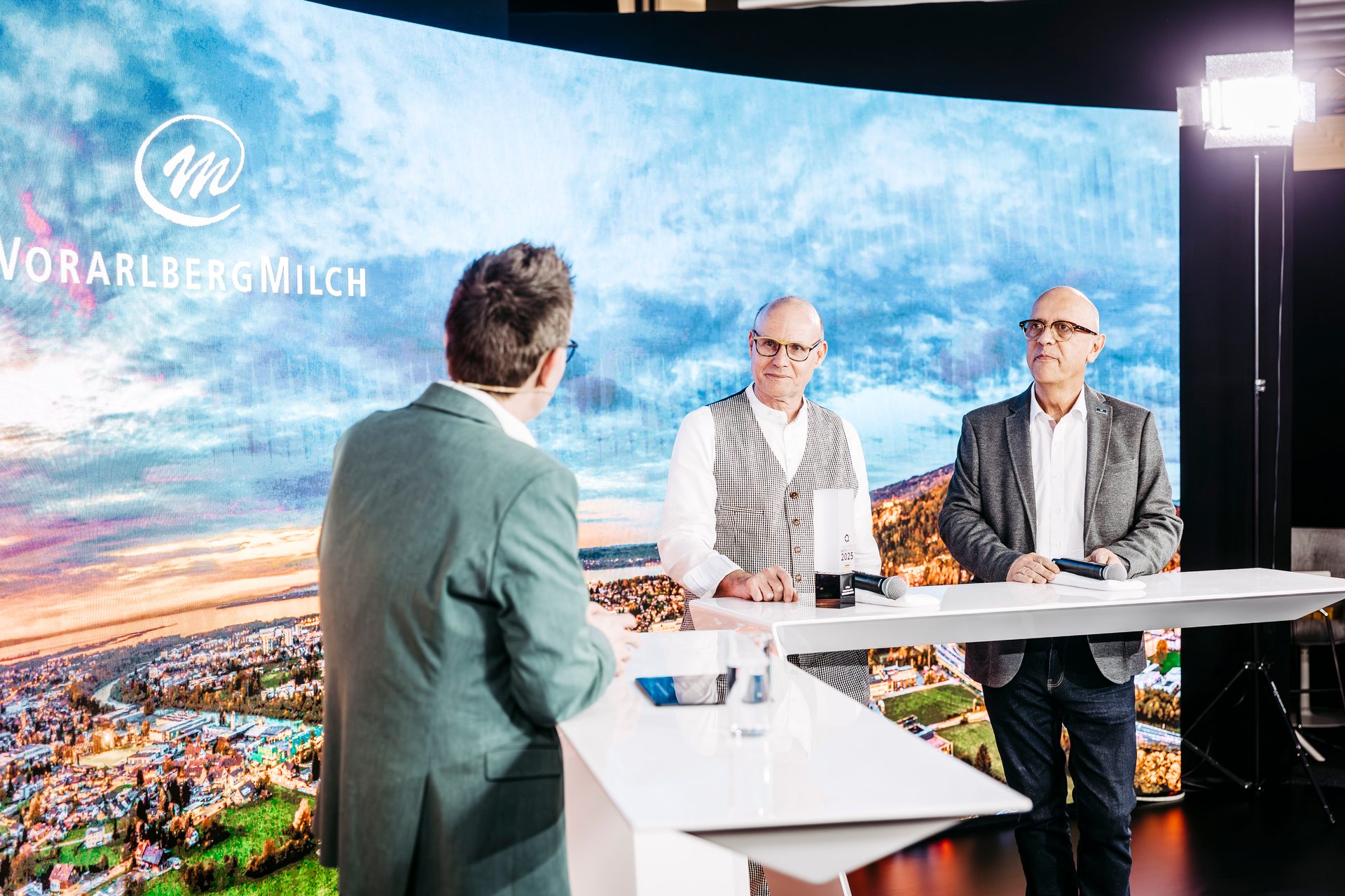 Three men presenting on a stage with a large screen displaying the Voralberg Milch logo and a landscape.
