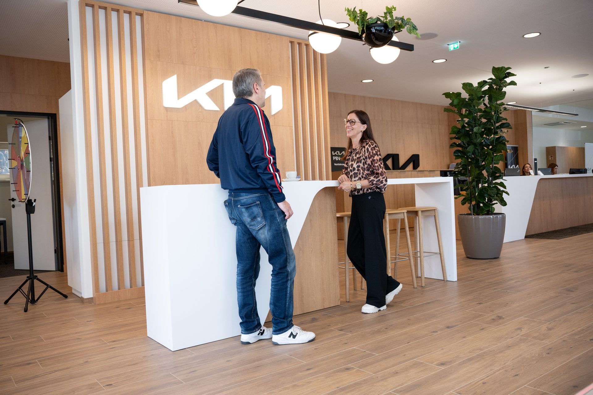 A man and a woman in casual attire converse at a white counter in a modern Kia dealership.