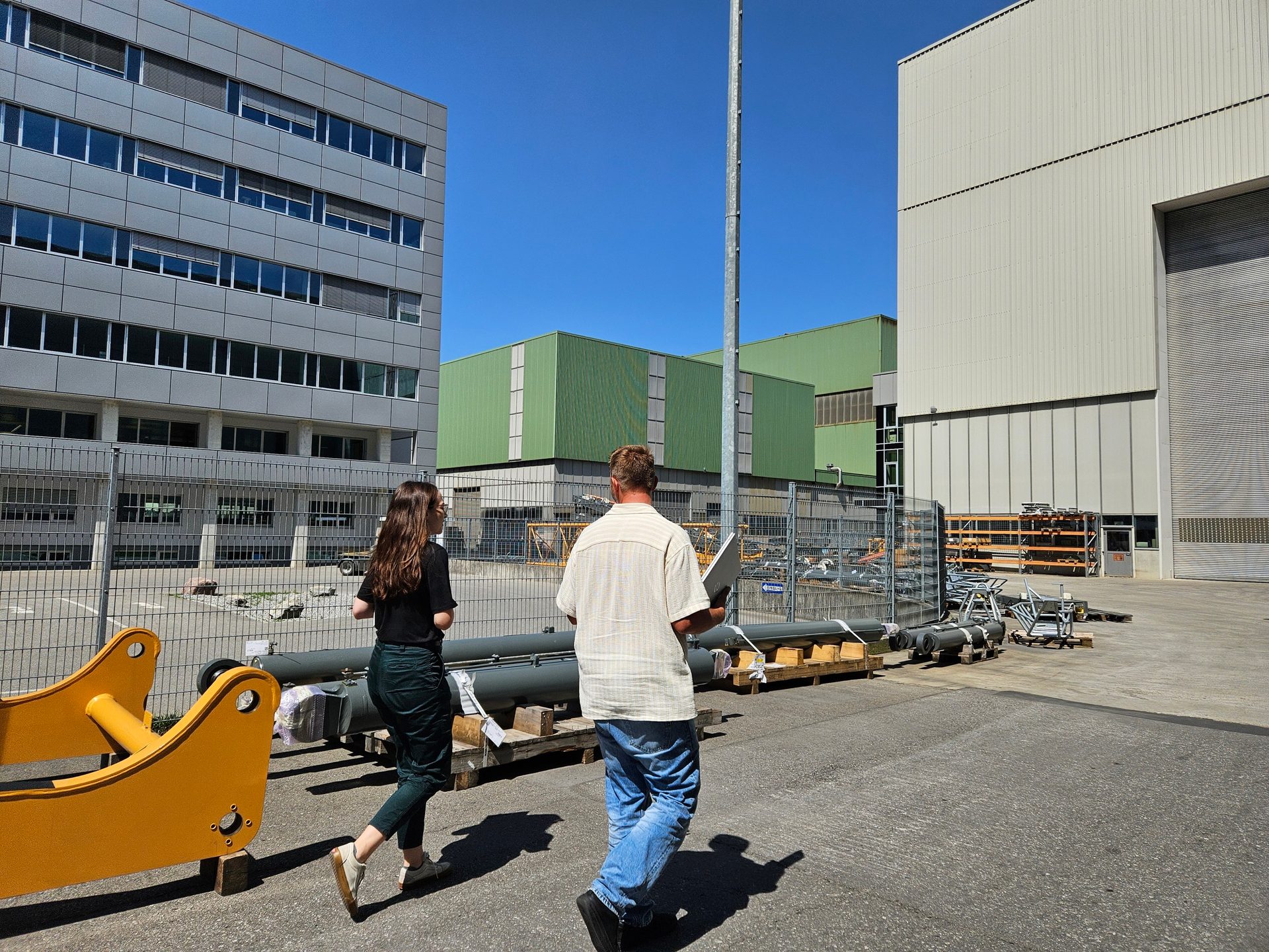 Two people walk past modern industrial buildings and stored equipment under a bright sky.