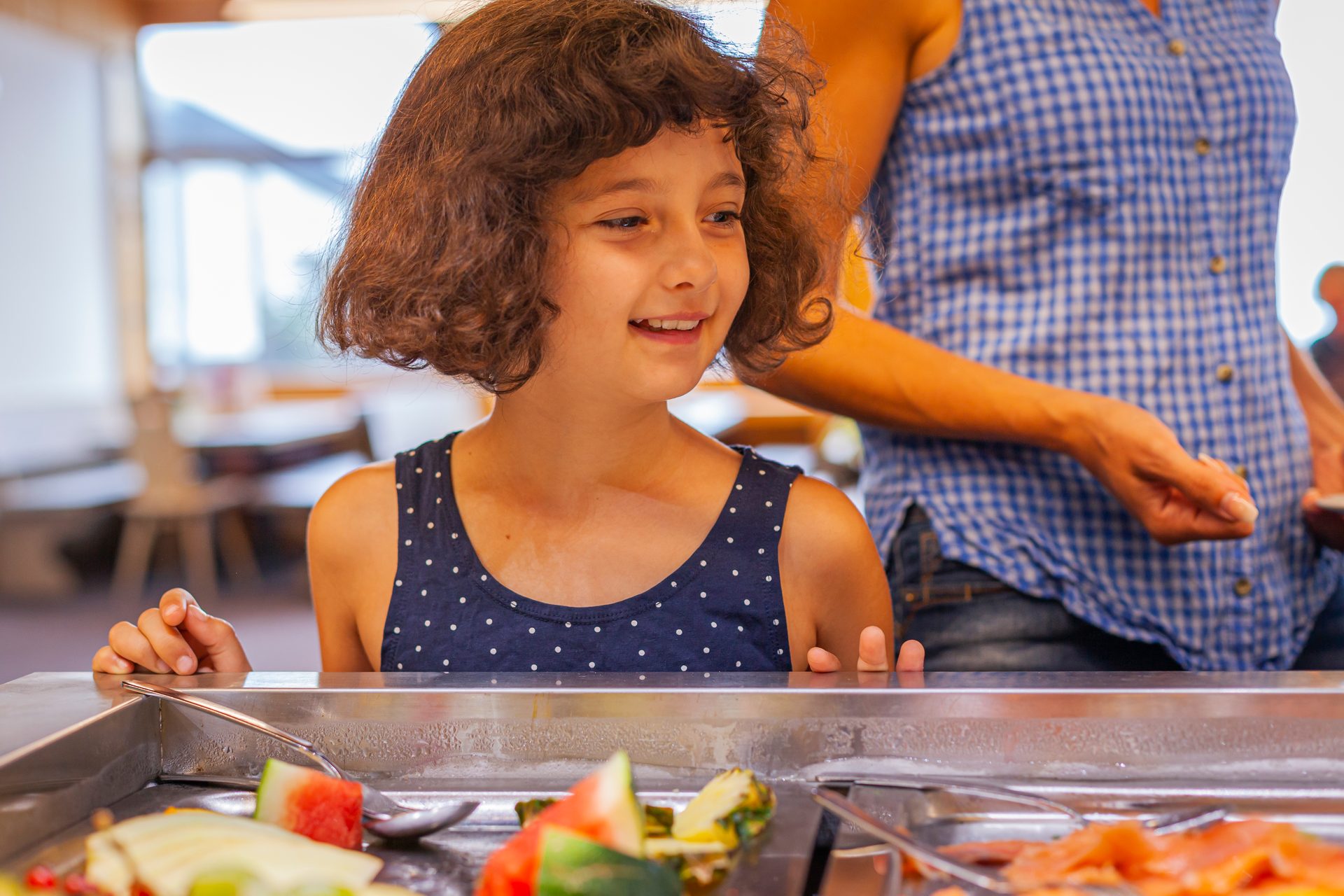 A smiling young girl with curly hair looks at a buffet line, with an adult behind her.
