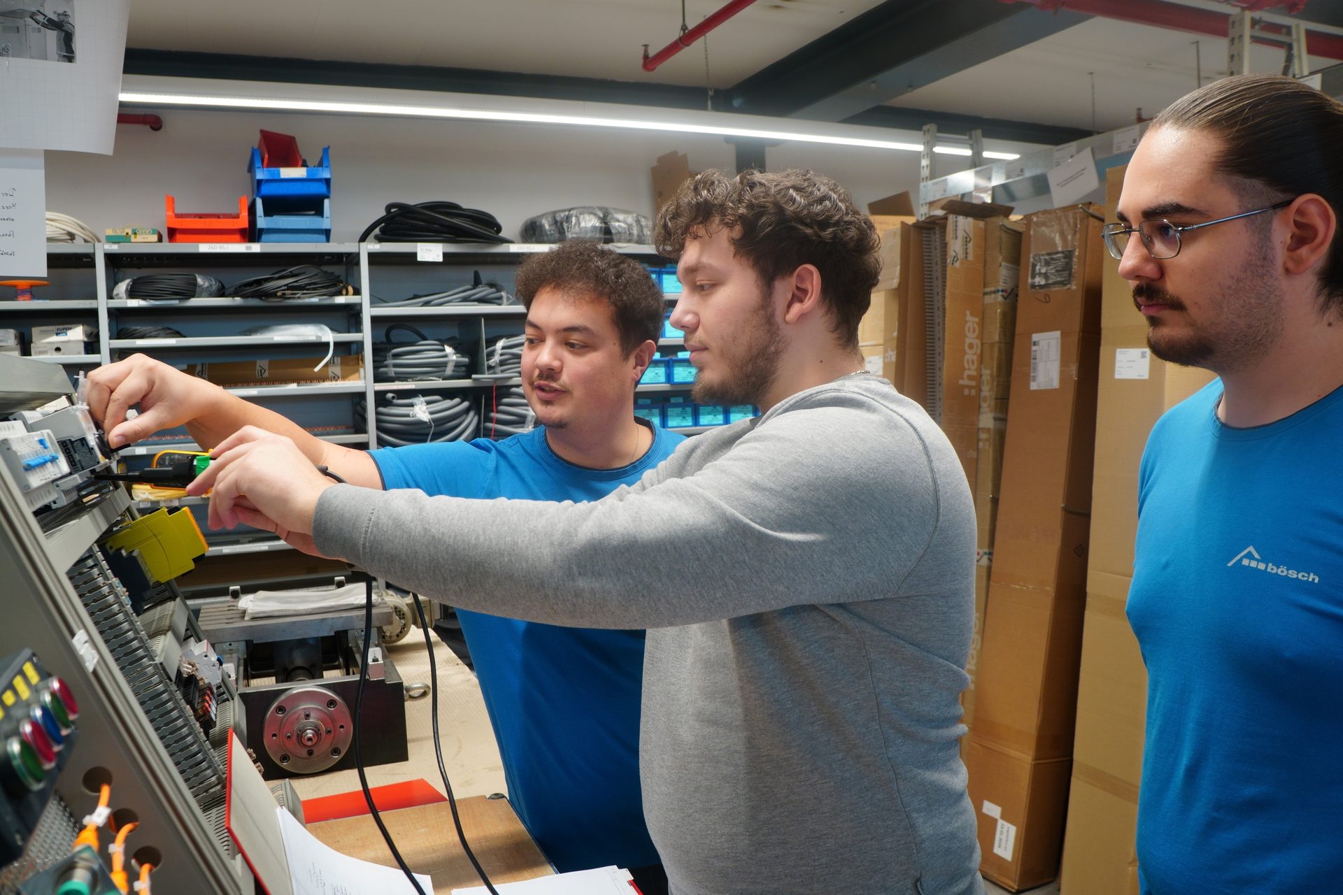 Three men in a workshop, two working on electrical equipment, one observing.