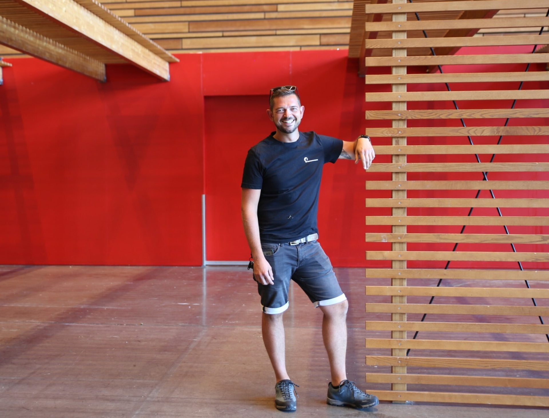 Young man smiling, leaning on a wooden screen in a modern room with red walls and wooden ceiling.