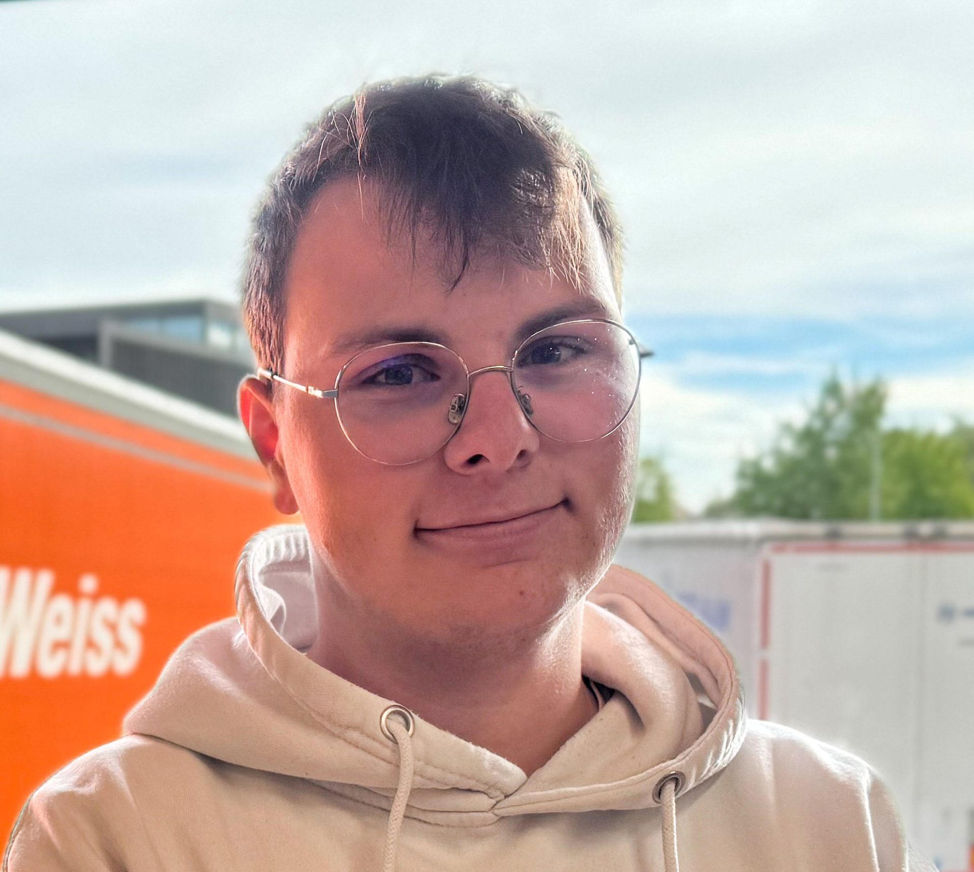A smiling person wearing round glasses and a light-colored hoodie, with a blurry outdoor background.