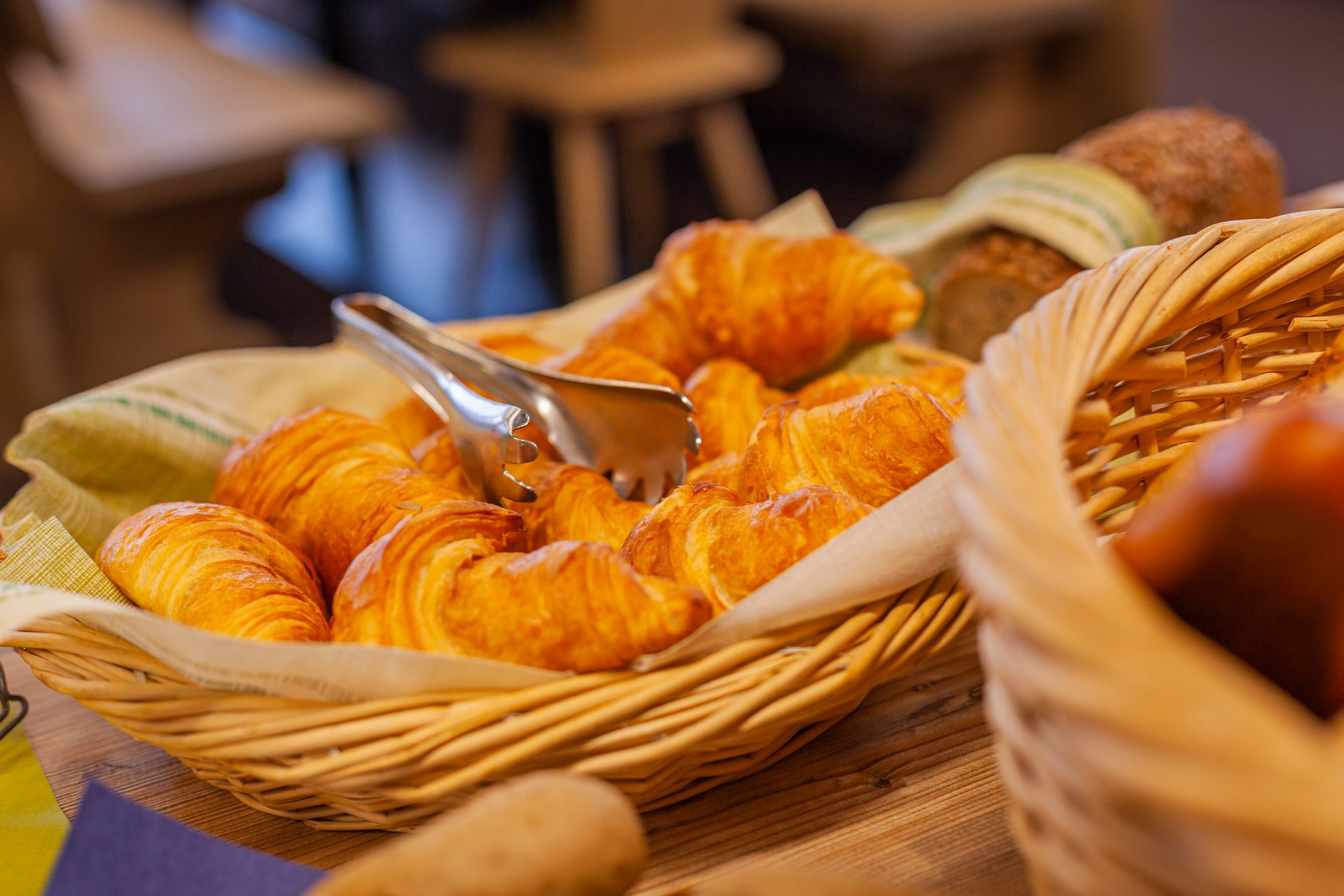 A woven basket filled with golden-brown croissants and metal serving tongs, ready for breakfast.