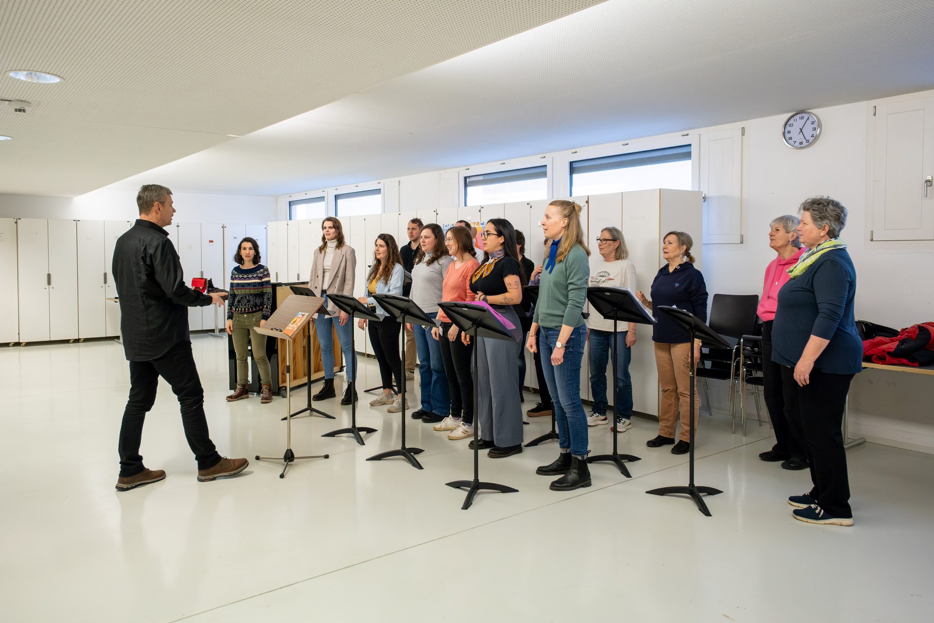 A conductor leads a diverse choir practicing in a bright, modern room with music stands.