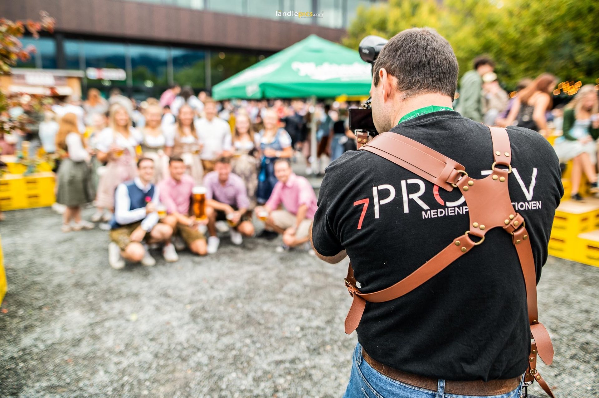 Photographer in a camera harness takes a photo of people with beer at an outdoor event.
